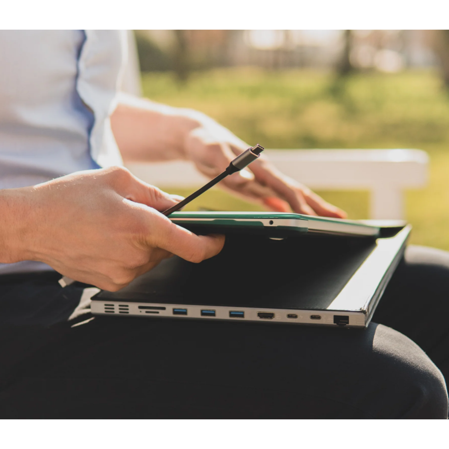 A person holding a laptop with a USB-C cable connected, sitting outdoors. The background is blurred.