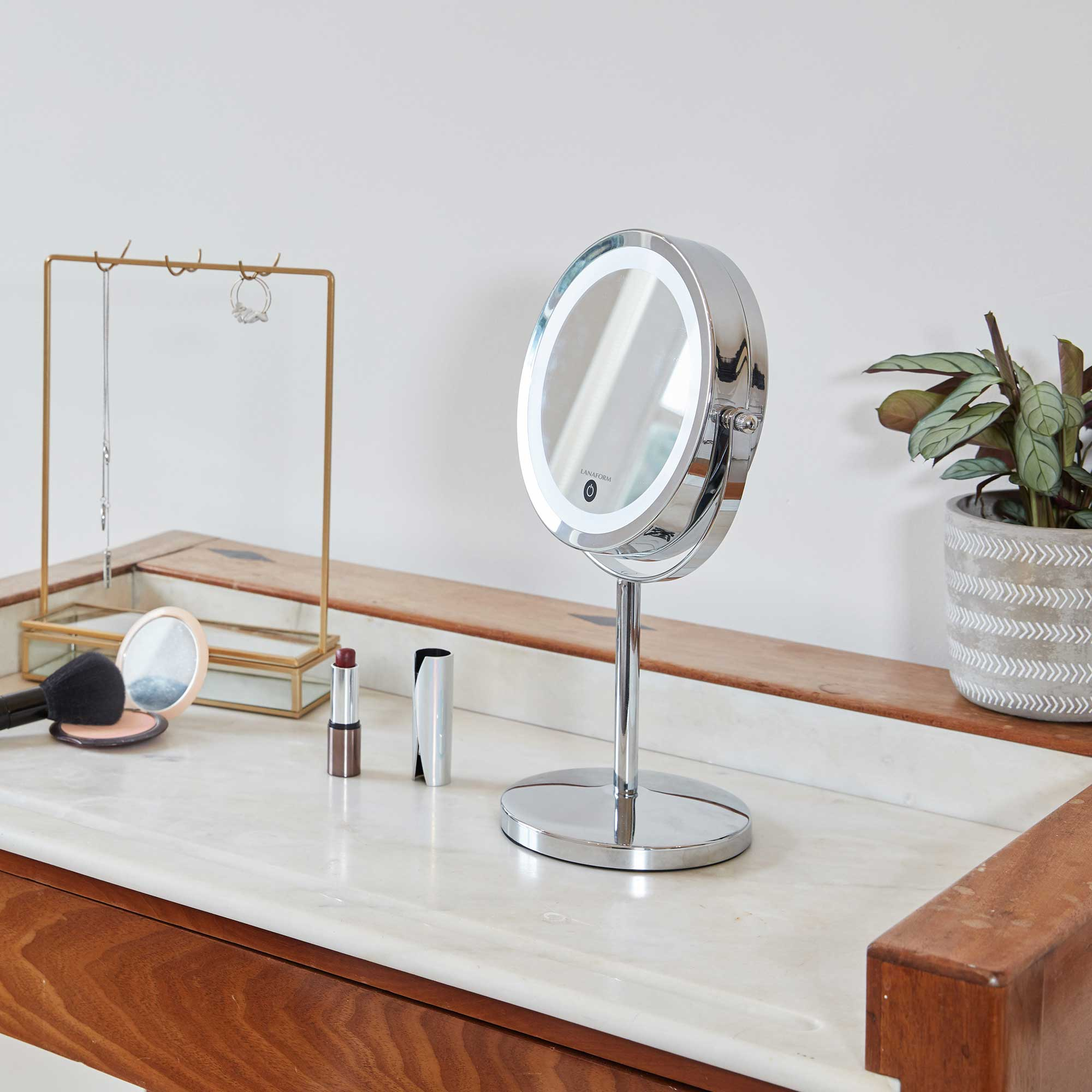 A vanity with a makeup mirror, lipstick, jewelry holder, and a potted plant. White wall backdrop.