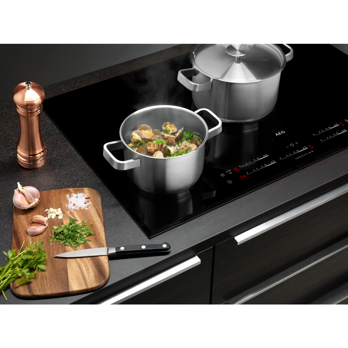 Escena de cocina con vitrocerámica negra, ollas e ingredientes en una tabla de madera.