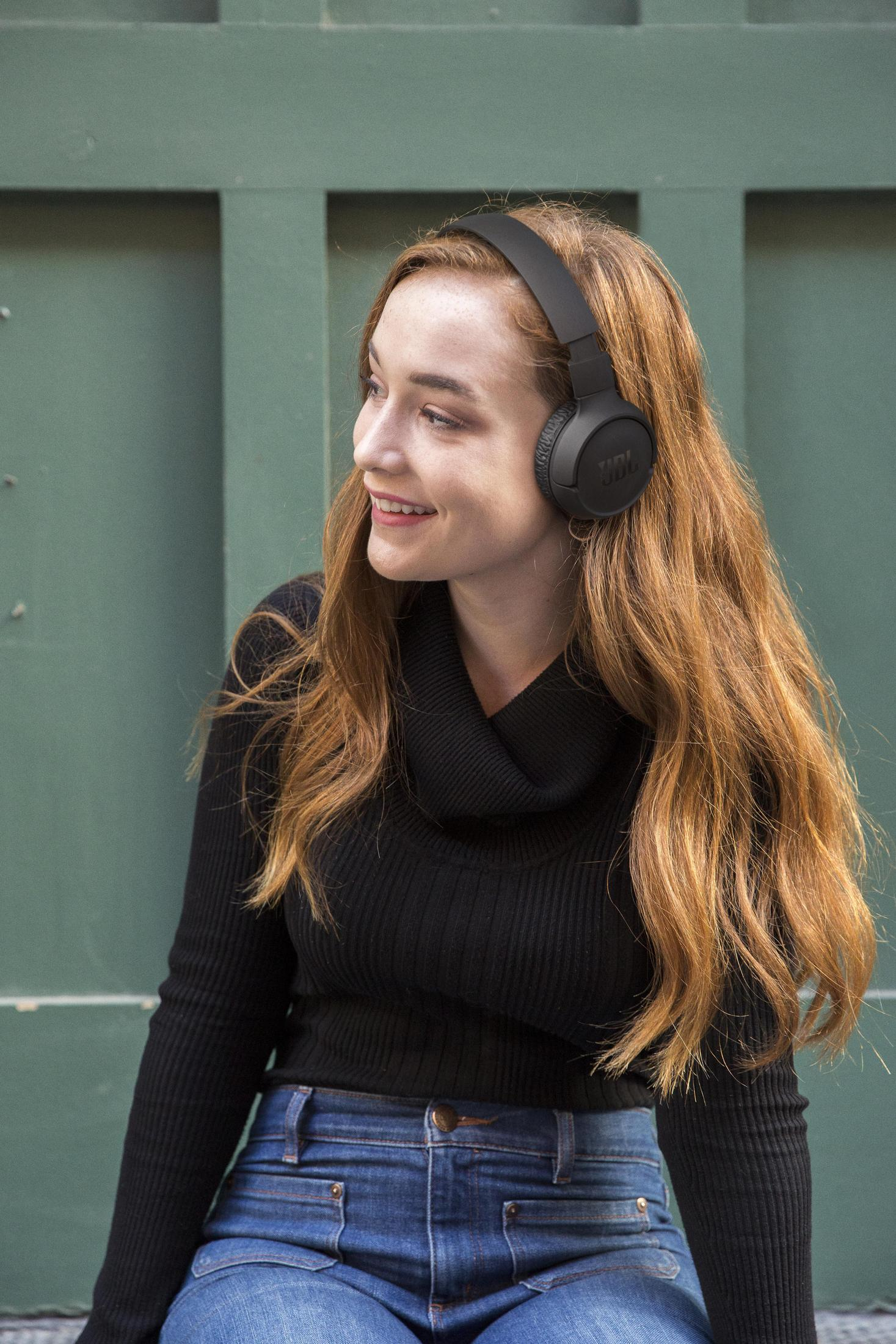 Uśmiechnięta kobieta z rudymi włosami nosi czarne słuchawki i czarny sweter, na tle zielonej ściany.