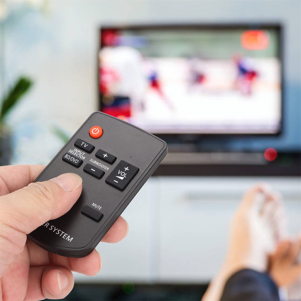Eine Hand, die eine Fernbedienung hält, im Hintergrund ist der Fernseher eingeschaltet. Die Fernbedienung ist schwarz mit Knöpfen.