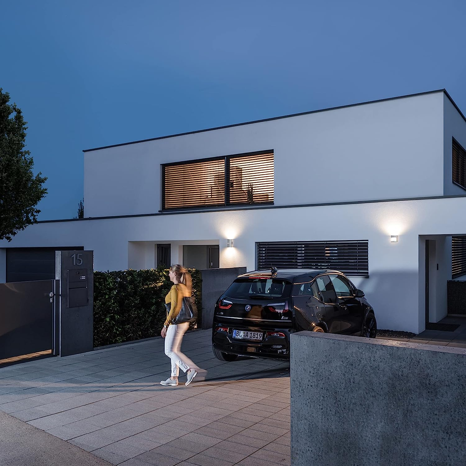 Frau geht auf ein schwarzes Auto zu, das vor einem modernen weißen Haus geparkt ist.
