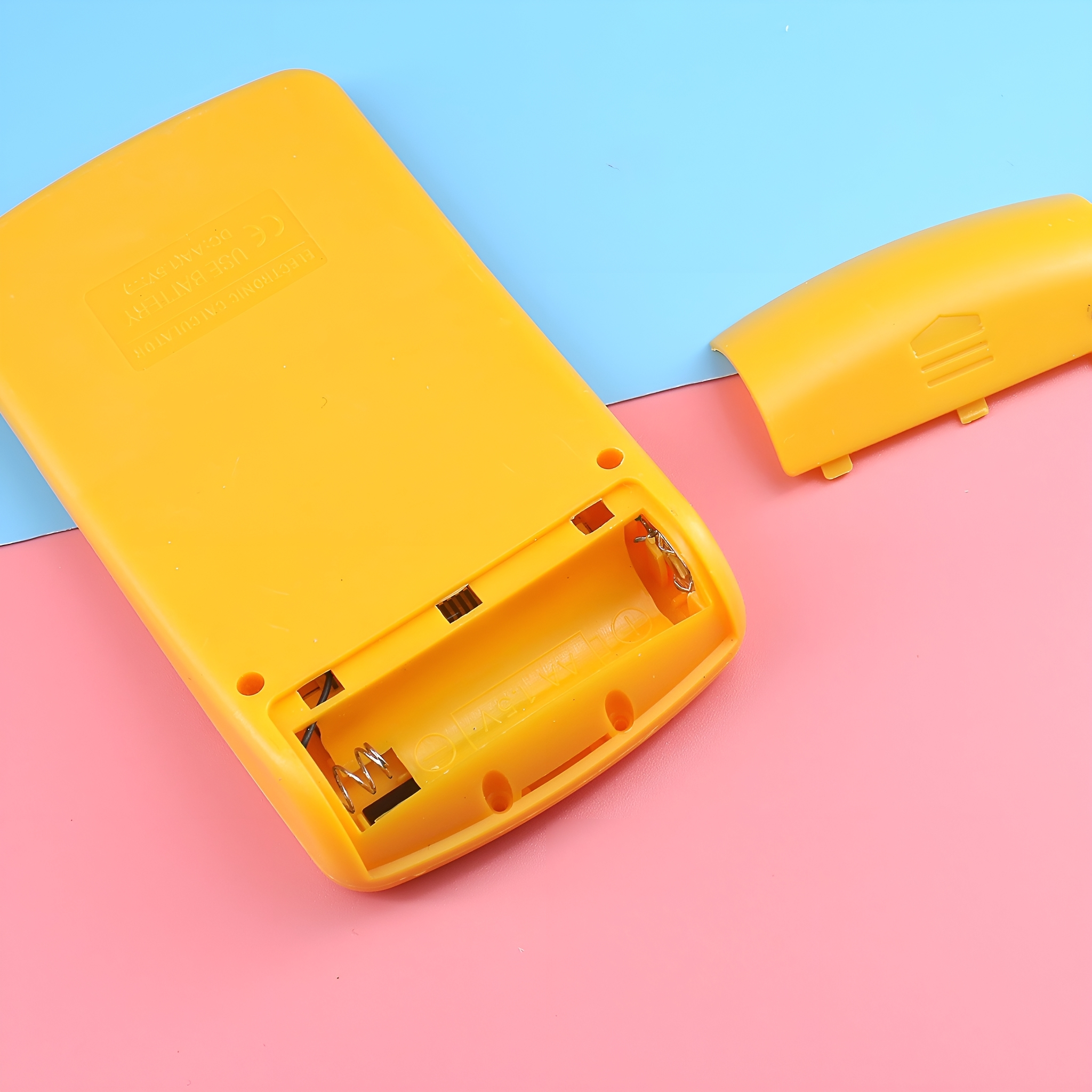 Gelbe Taschenrechnerhülle mit offenem Batteriefach, rosa und blauer Hintergrund.