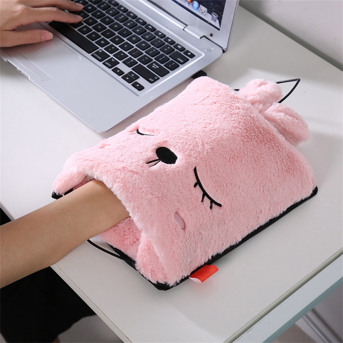 Eine rosa, flauschige Handwärmer mit Gesicht, eine Hand darin und ein Laptop daneben.