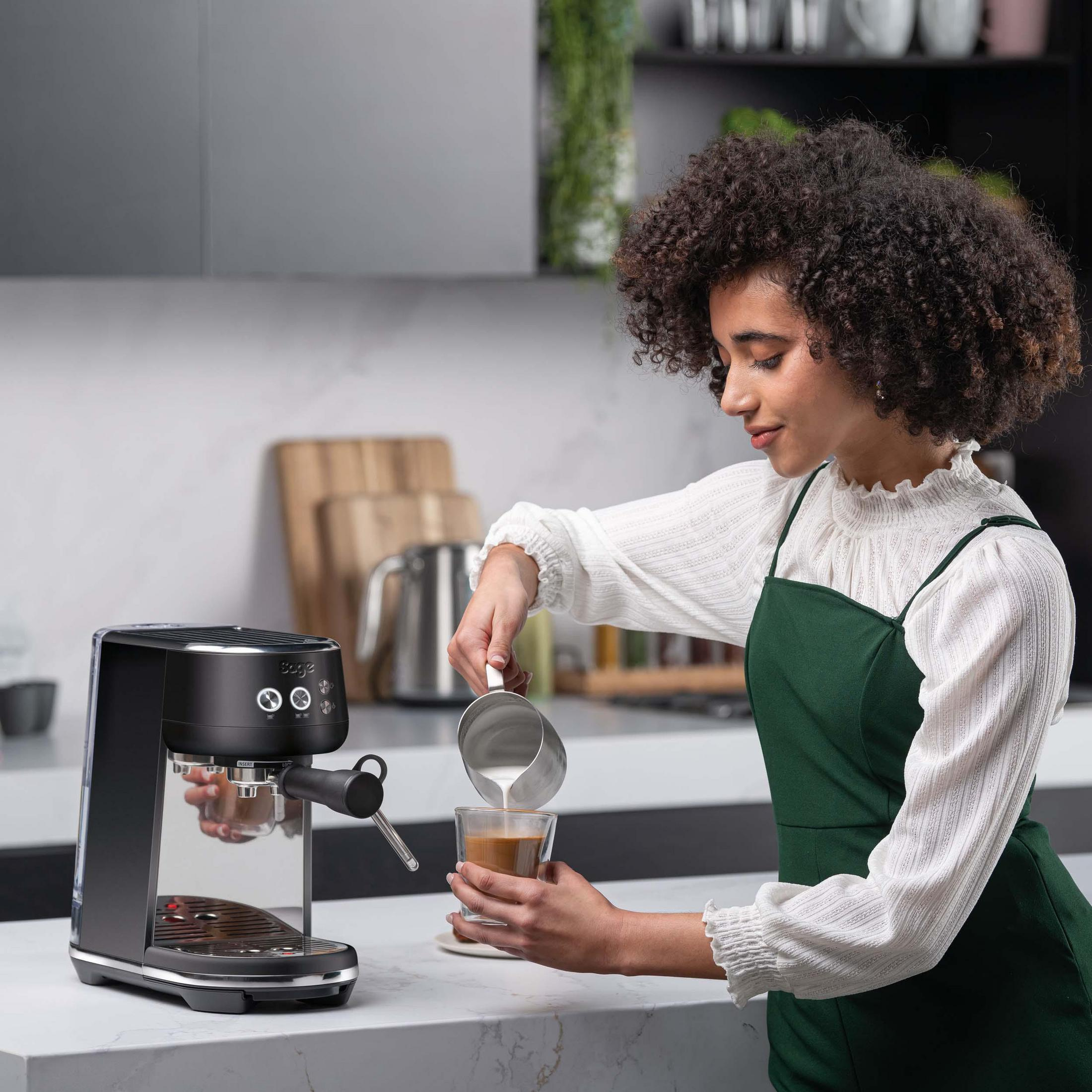 Kobieta wlewa mleko do kawy ze srebrnego dzbanka obok czarnego ekspresu do kawy.