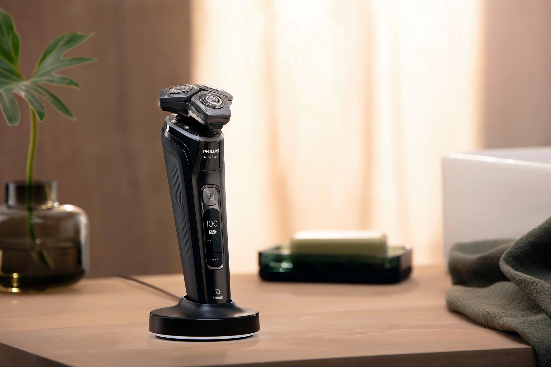 Czarna golarka Philips na stacji ładującej na drewnianej powierzchni z zieloną rośliną i mydelniczką.