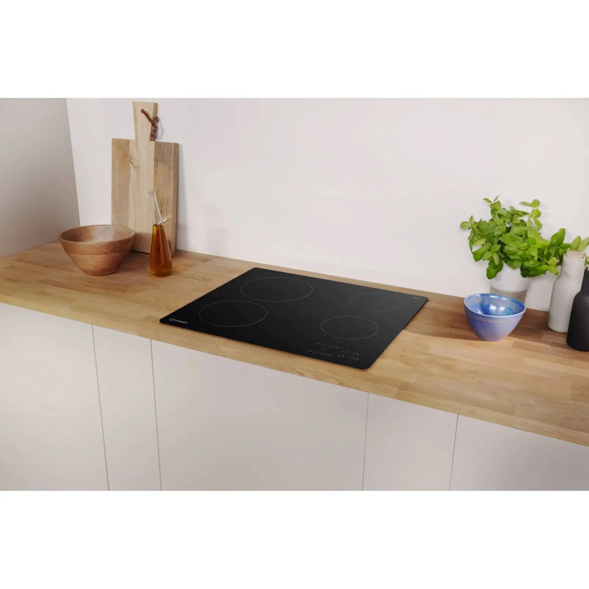Placa negra en encimera de madera. Tabla de cortar, cuenco, botella de aceite y plantas al fondo.