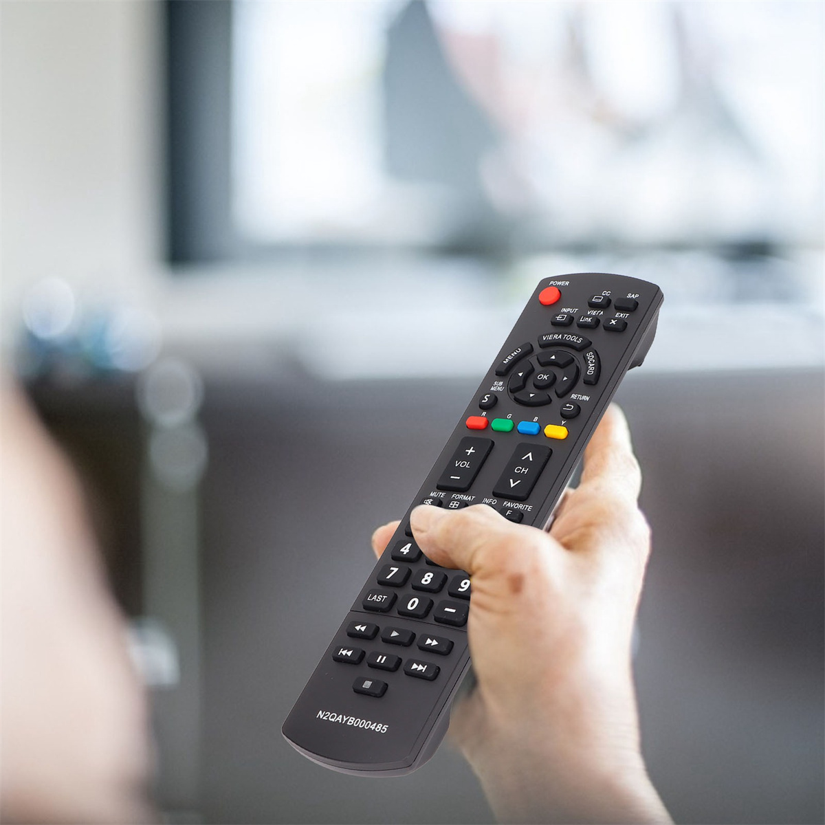Eine Hand, die eine schwarze Fernbedienung mit vielen Tasten hält. Im Hintergrund ist ein verschwommener Fernseher zu sehen.
