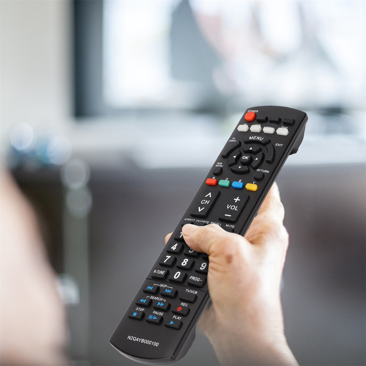 Eine Person hält eine schwarze Fernbedienung mit verschiedenen Knöpfen. Im Hintergrund ein Fernseher.