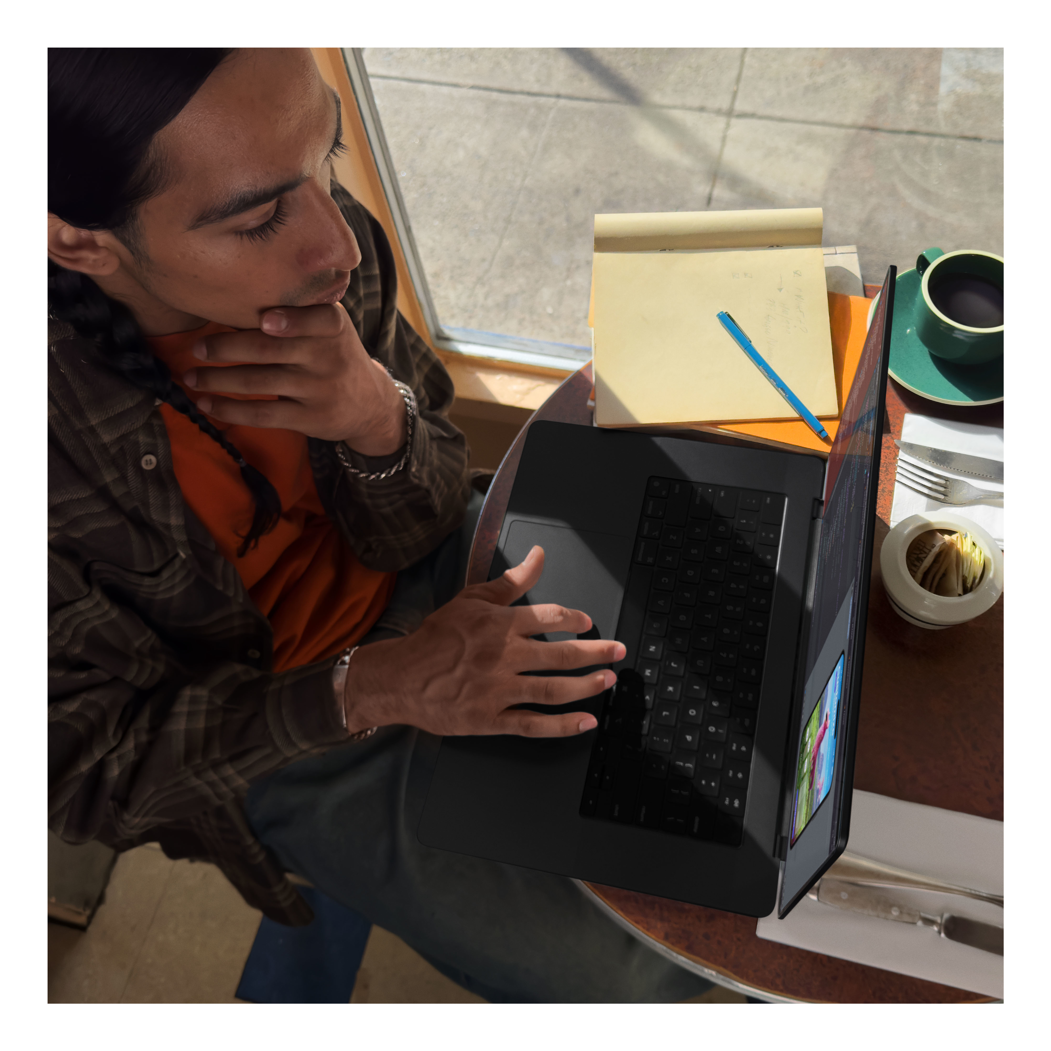 Ein Mann mit langen Haaren arbeitet in einem Café an einem Laptop. Eine Tasse und ein Notizblock sind vorhanden.