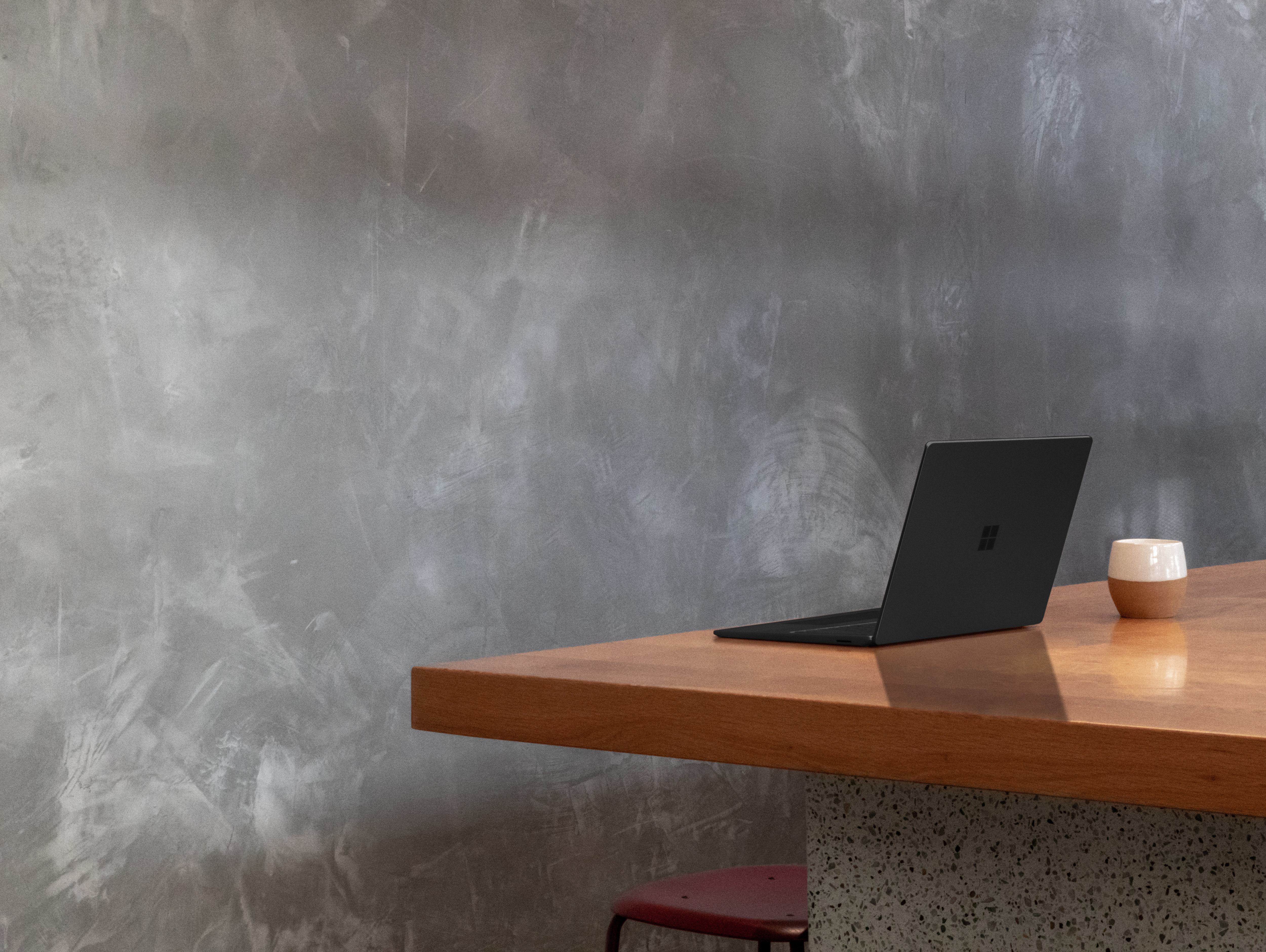 Ein schwarzer Laptop ist auf einem Holztisch geöffnet. Der Hintergrund ist eine graue Wand und eine kleine Tasse.