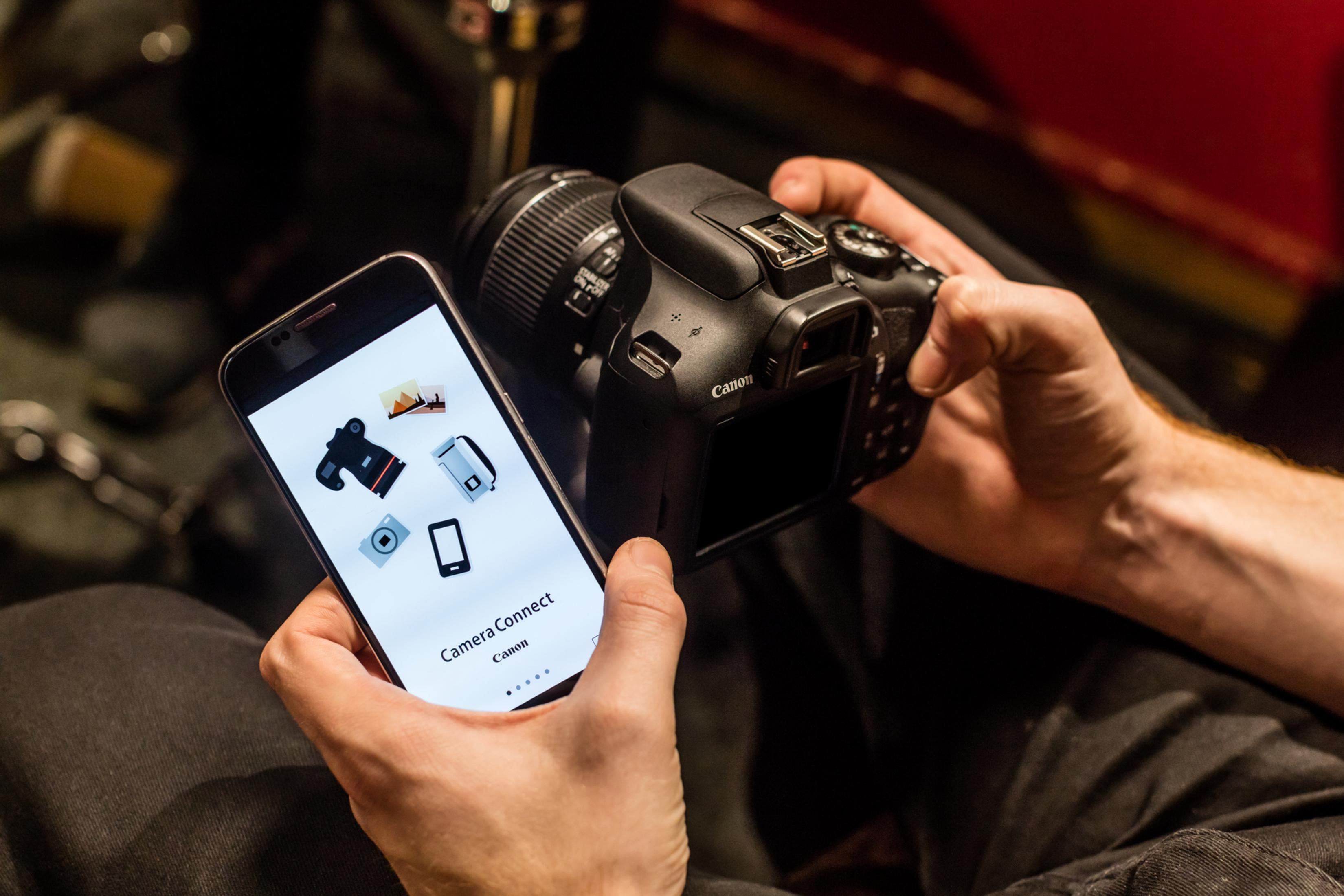 Osoba trzymająca telefon i aparat. Telefon pokazuje aplikację aparatu. Czarne tło.