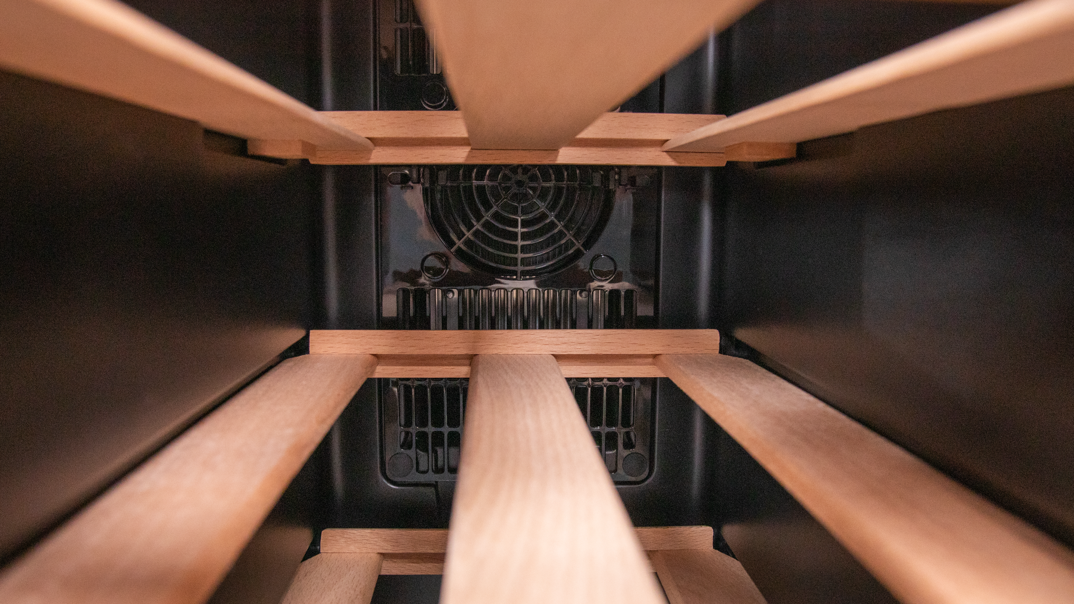 Wewnątrz chłodziarki do wina: drewniane półki i wentylator. Tło jest ciemne.