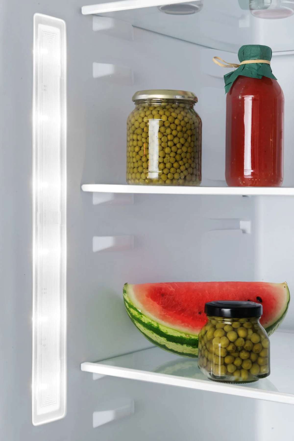 Wnętrze lodówki z żywnością. Zawiera słoiki z groszkiem, butelkę sosu i arbuza.