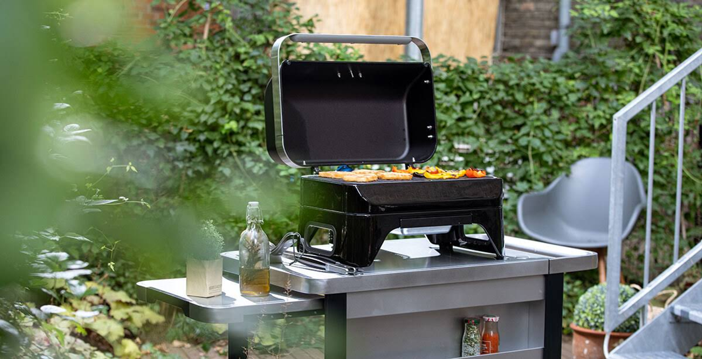 Een draagbare zwarte grill op een tafel. Er worden groenten op gekookt. Groene struiken en een grijze stoel op de achtergrond.