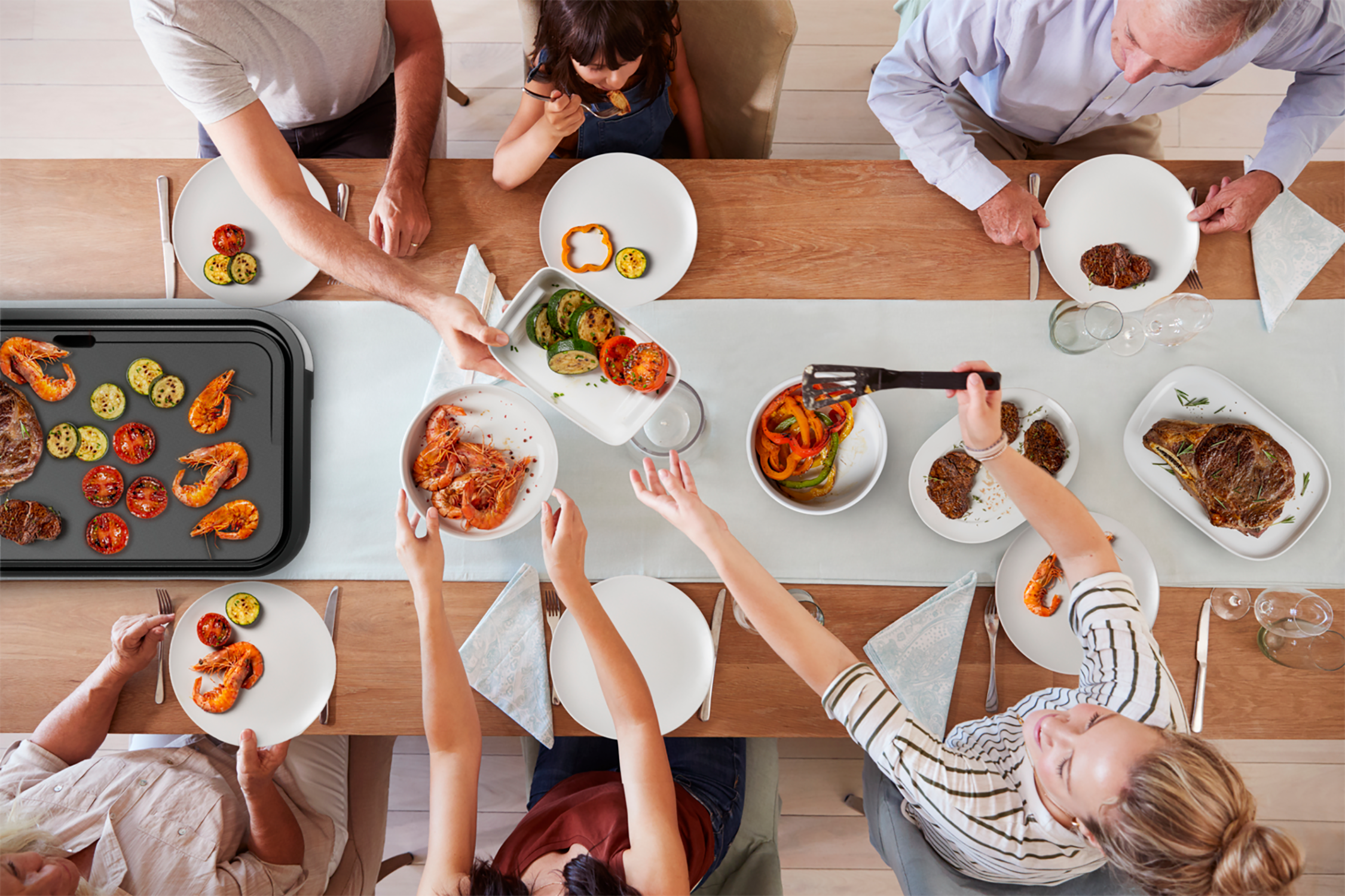 Widok z góry na rodzinę przy stole z jedzeniem, talerzami i grillem pośrodku.