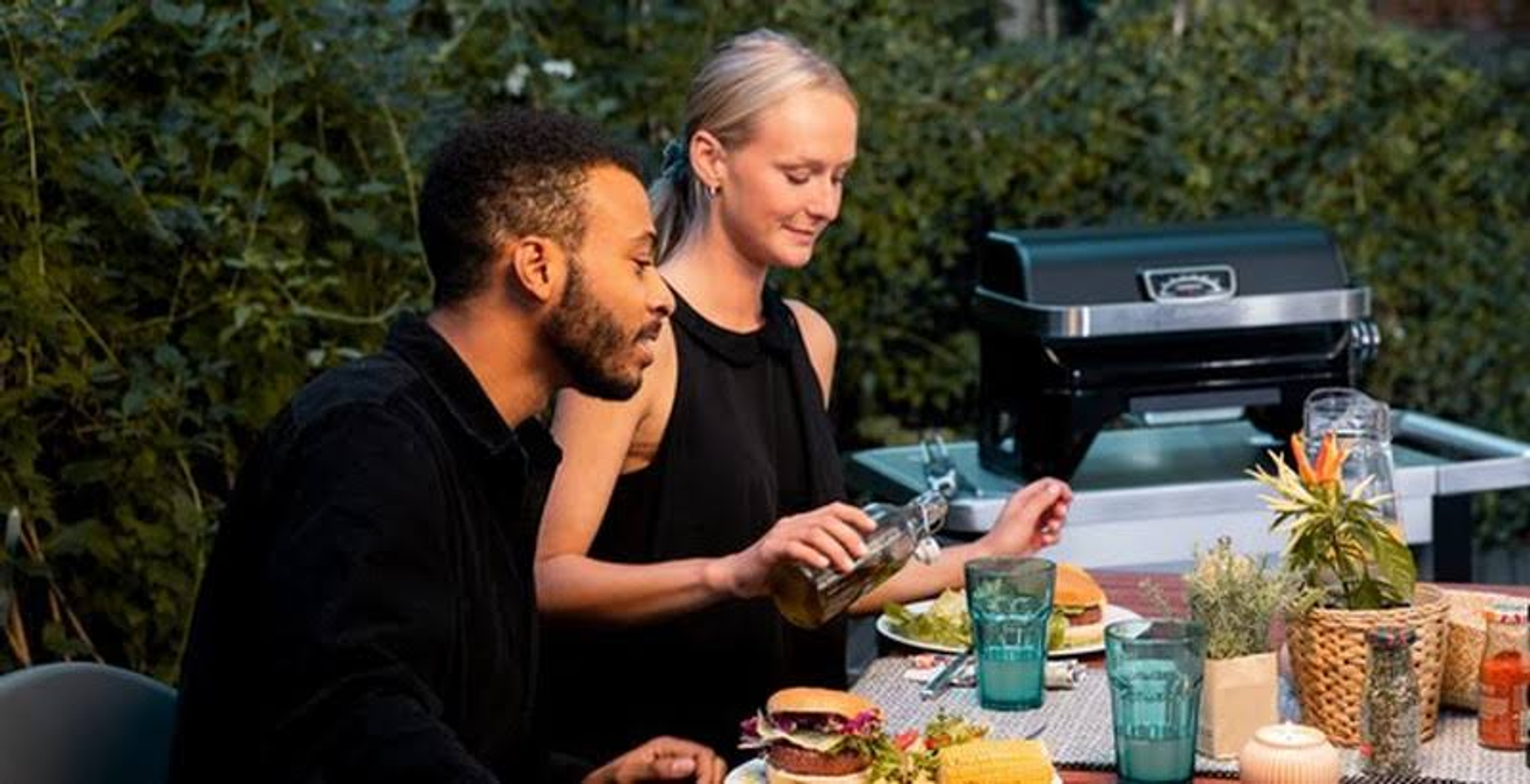 Een stel aan een tafel met eten en een grill op de achtergrond, genietend van een maaltijd in de buitenlucht.