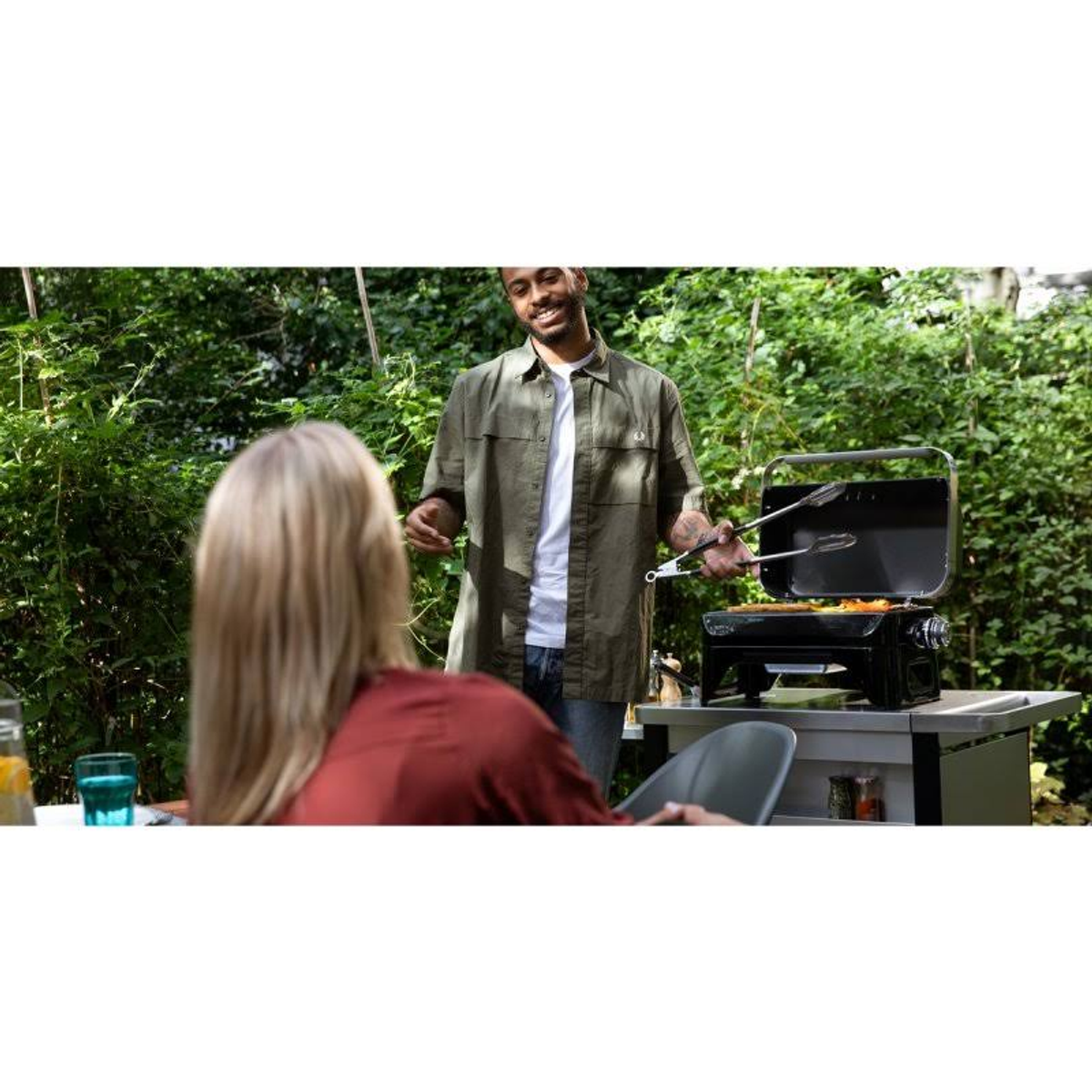 Mann grillt draußen Essen, während eine Frau an einem Tisch sitzt, grüner Hintergrund.