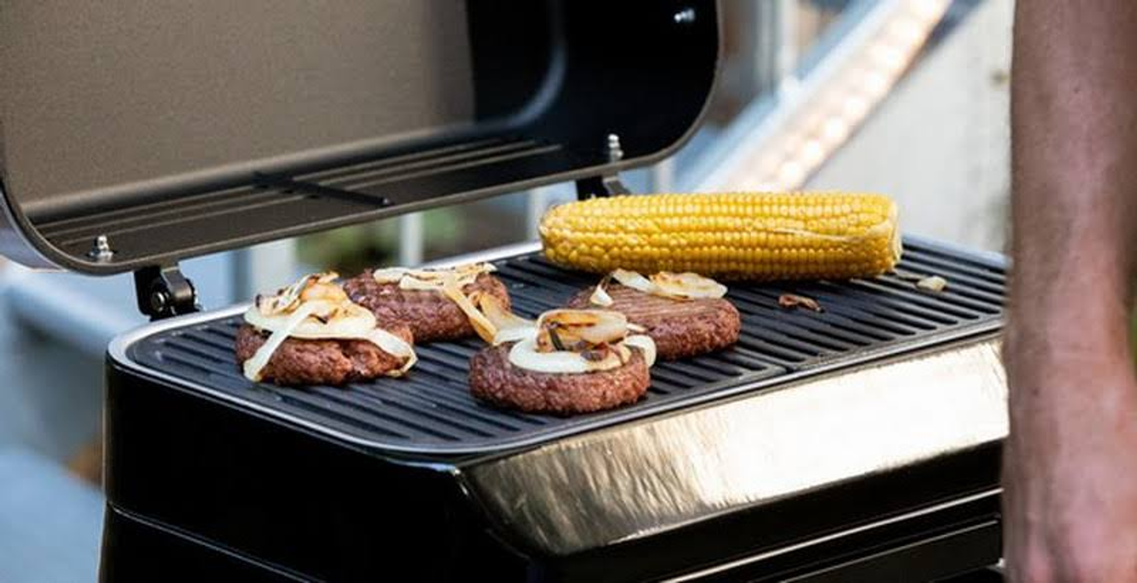 Parrilla con hamburguesas, cebollas y mazorcas de maíz, parrilla negra, fondo borroso.