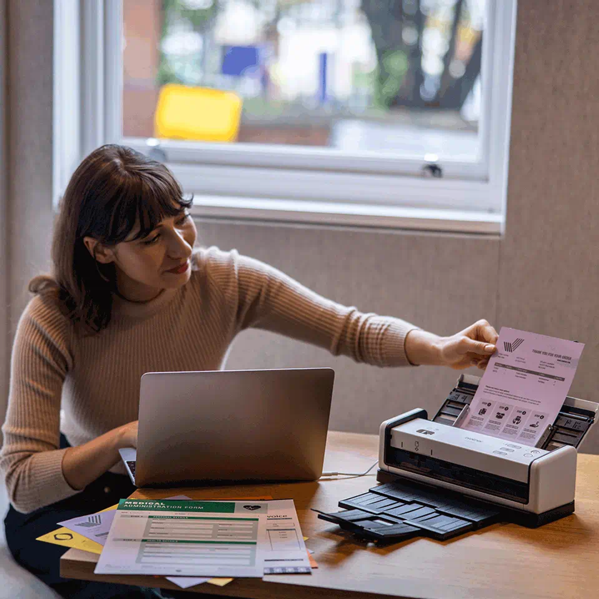 Kobieta skanuje dokument z laptopem i skanerem. Papiery na stole, okno w tle.
