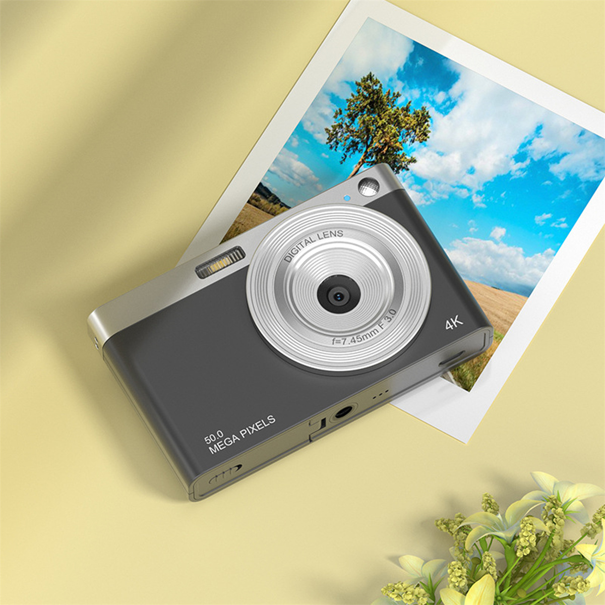 Eine Digitalkamera mit silbernem Objektiv und grauem Gehäuse, neben einem Foto und gelber Fläche.