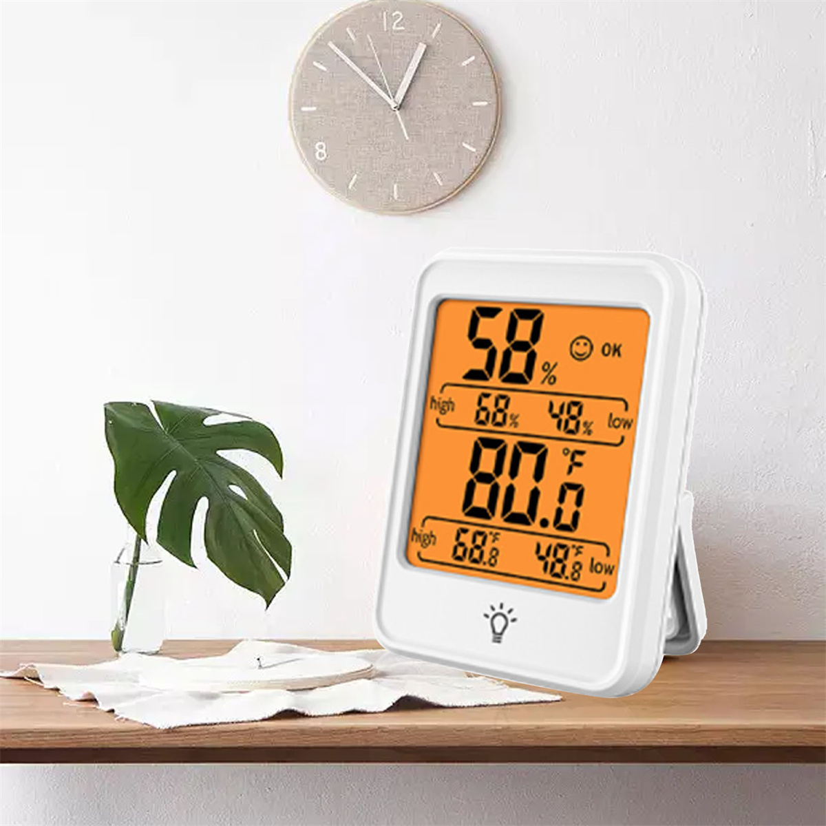 Weißes digitales Thermometer, das Temperatur und Luftfeuchtigkeit anzeigt, mit Uhr im Hintergrund.