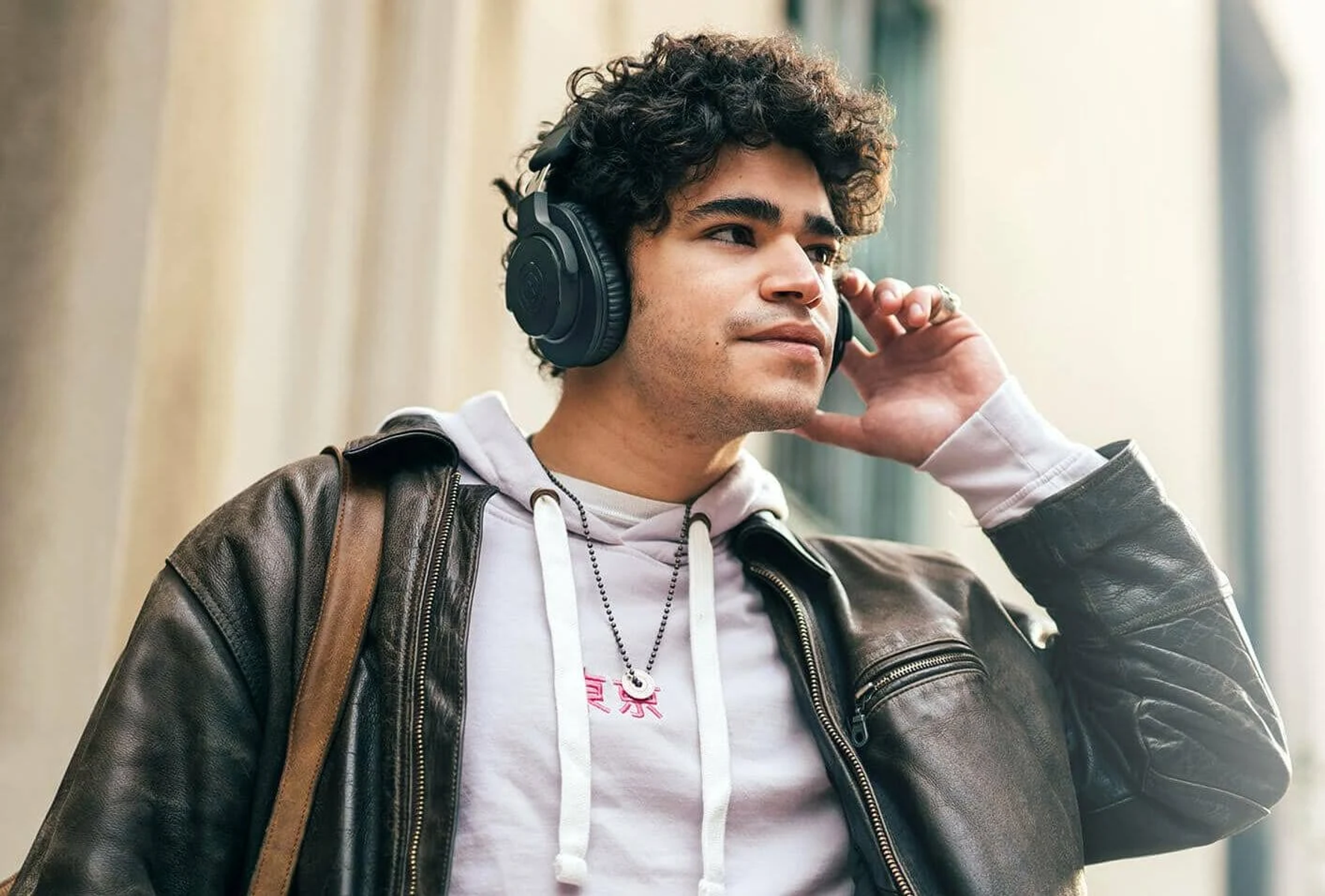 Osoba z kręconymi włosami ma na sobie słuchawki, bluzę z kapturem i skórzaną kurtkę.