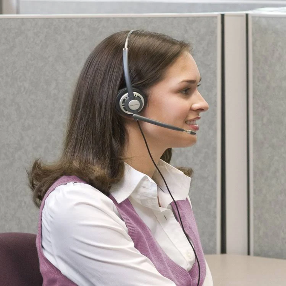 Kobieta w zestawie słuchawkowym uśmiecha się, siedząc w pobliżu szarej ściany kabiny.