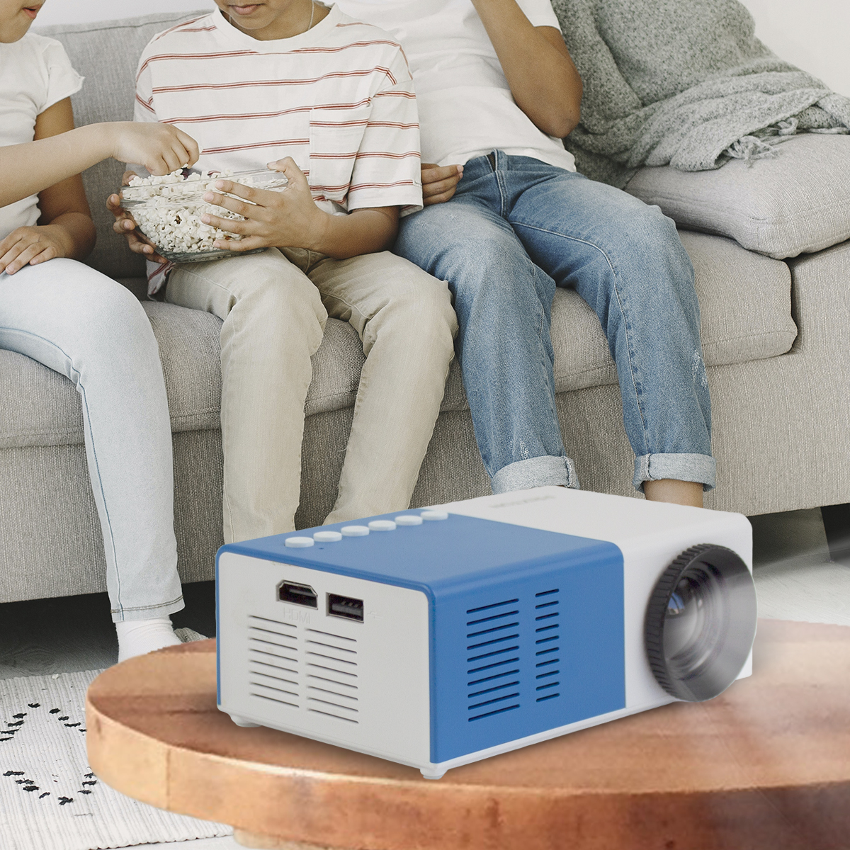Een witte en blauwe projector op een tafel. Drie mensen zitten op een bank op de achtergrond.