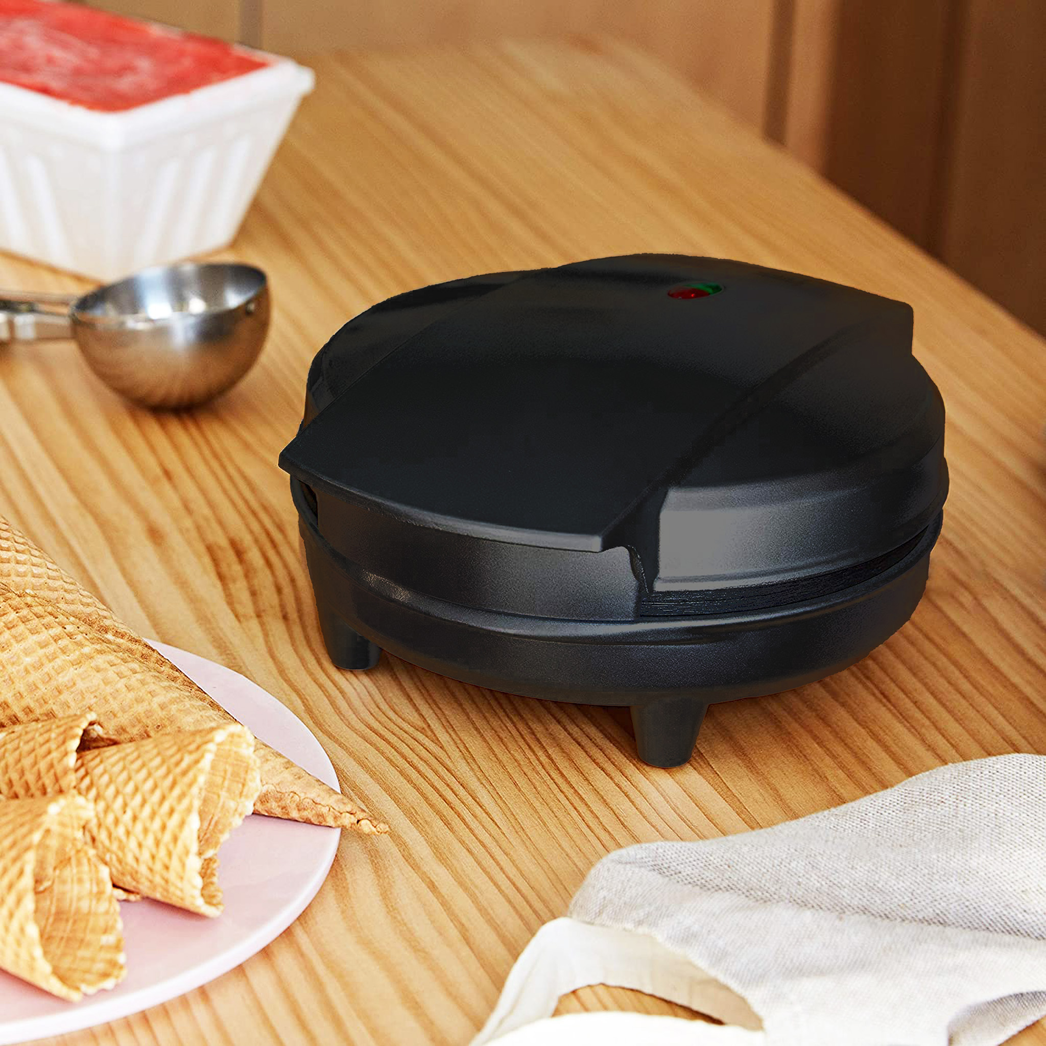 A black waffle maker on a wooden table with waffle cones and other items.