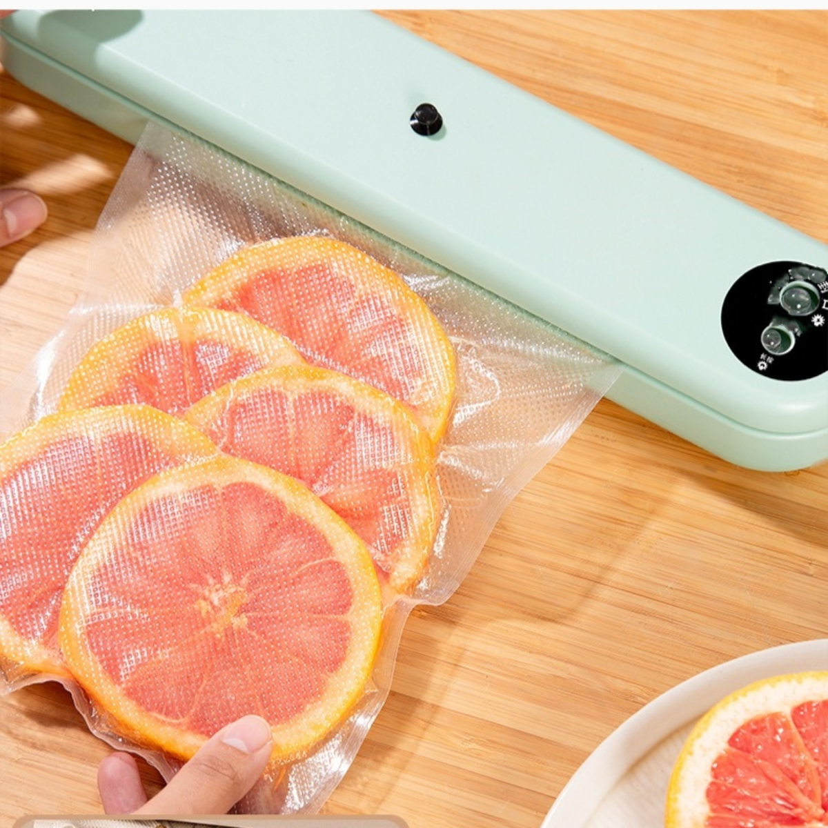 Ein Vakuumiergerät mit Grapefruit-Scheiben in einem Plastikbeutel auf einem Holztisch. Eine Hand ist auf dem Beutel.