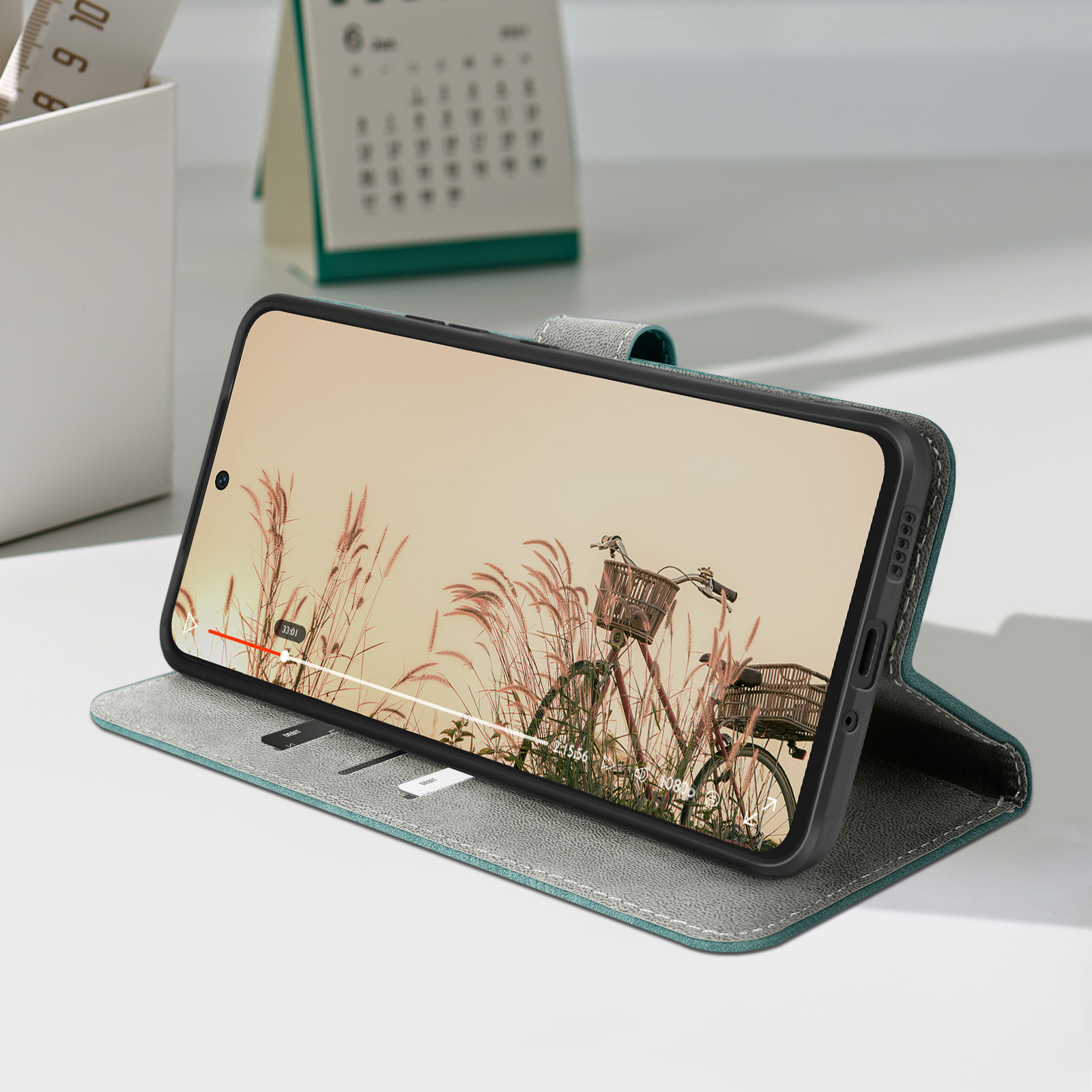 Telefono in custodia mostra video di una bici in un campo. Calendario e portapenne sullo sfondo.