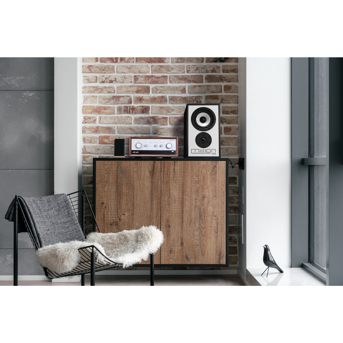Living room with a cabinet, speakers, a chair, and a brick wall. Bird statue on window sill.