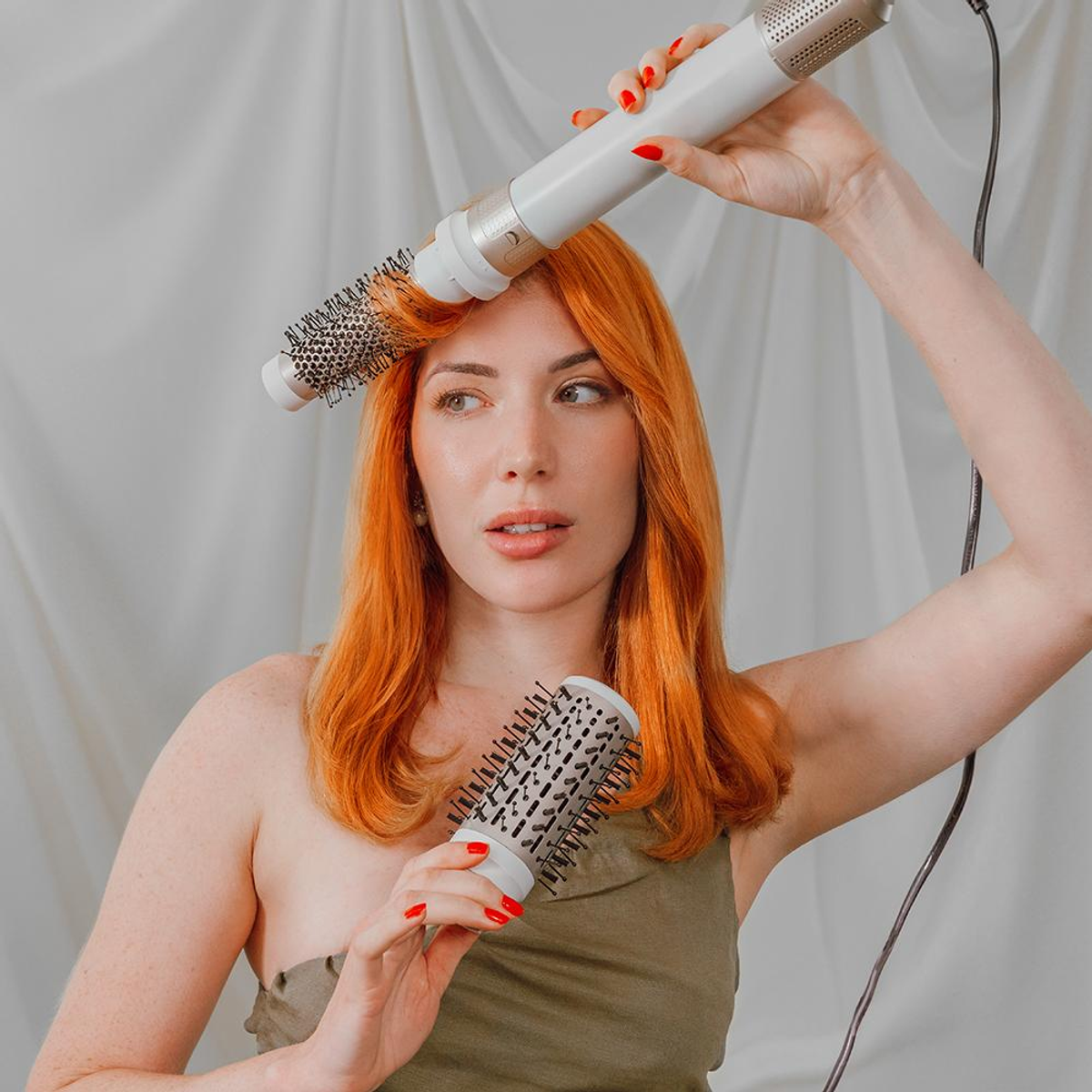 Woman holding two hair styling tools, red hair and nails. White background.