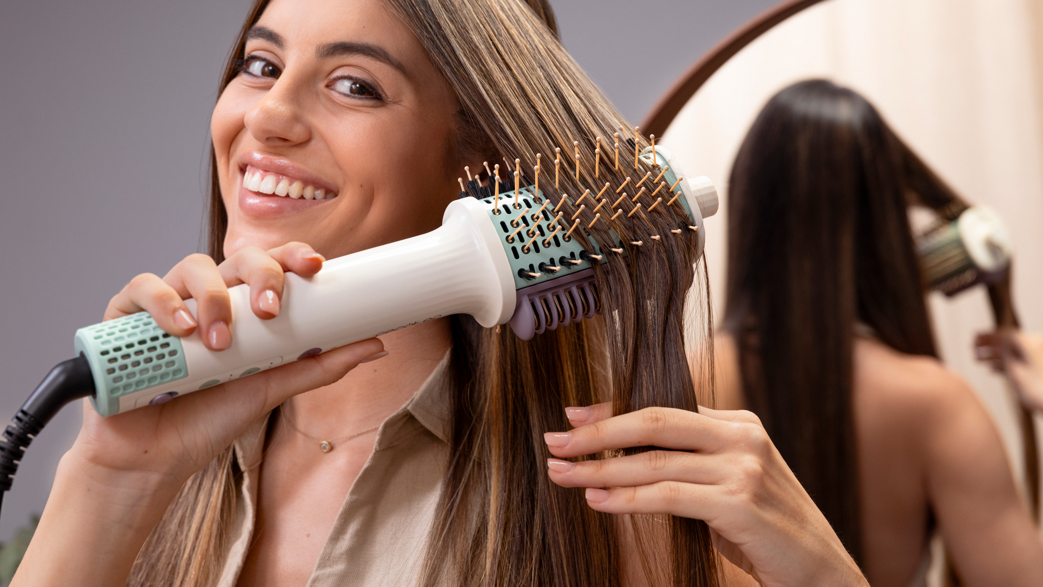 Donna che si acconcia i capelli con una spazzola. Sorride, guarda la fotocamera. Specchio sullo sfondo.
