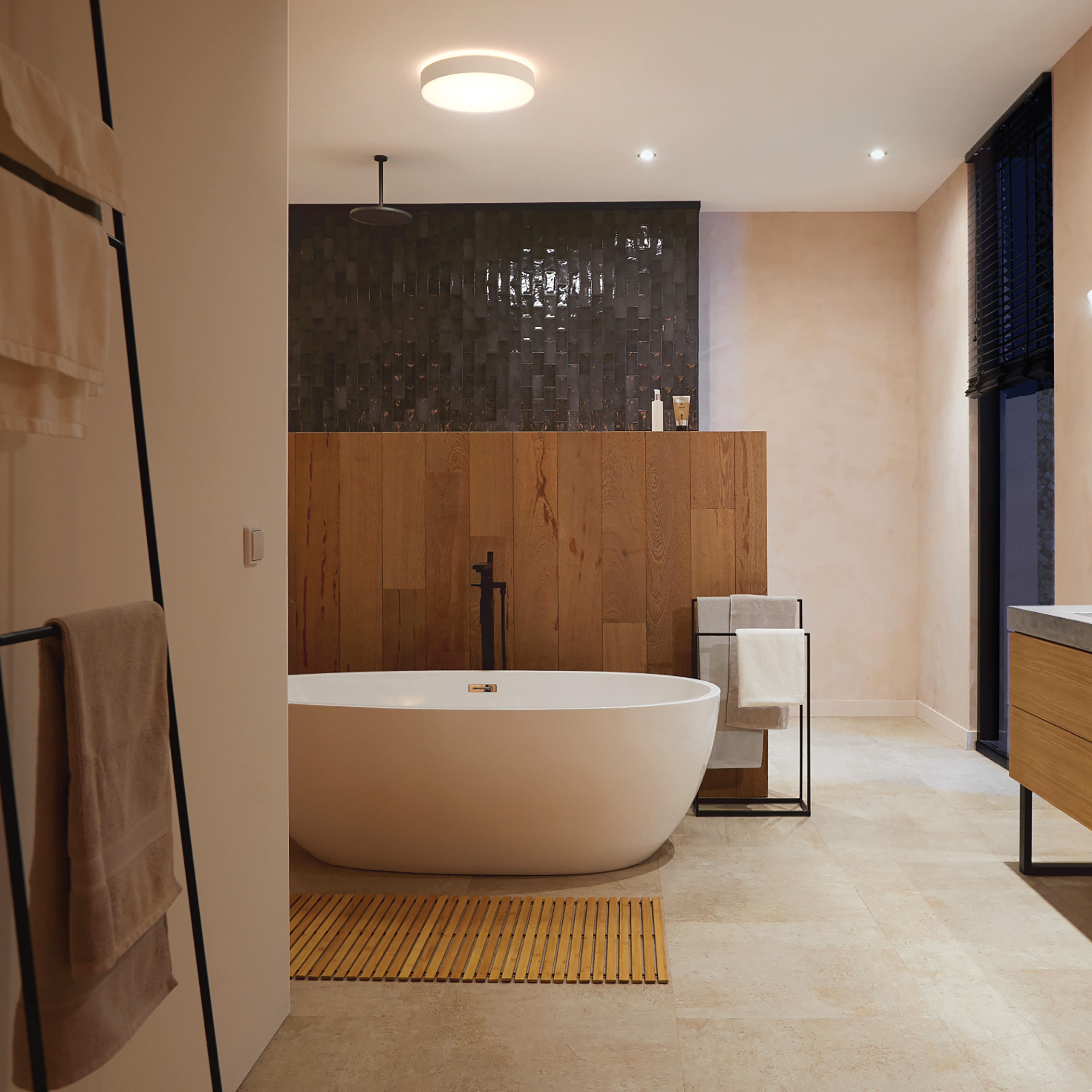Luksusowa łazienka. Biała wanna, drewniana ściana, czarne płytki i beżowa podłoga. Oświetlenie sufitowe.