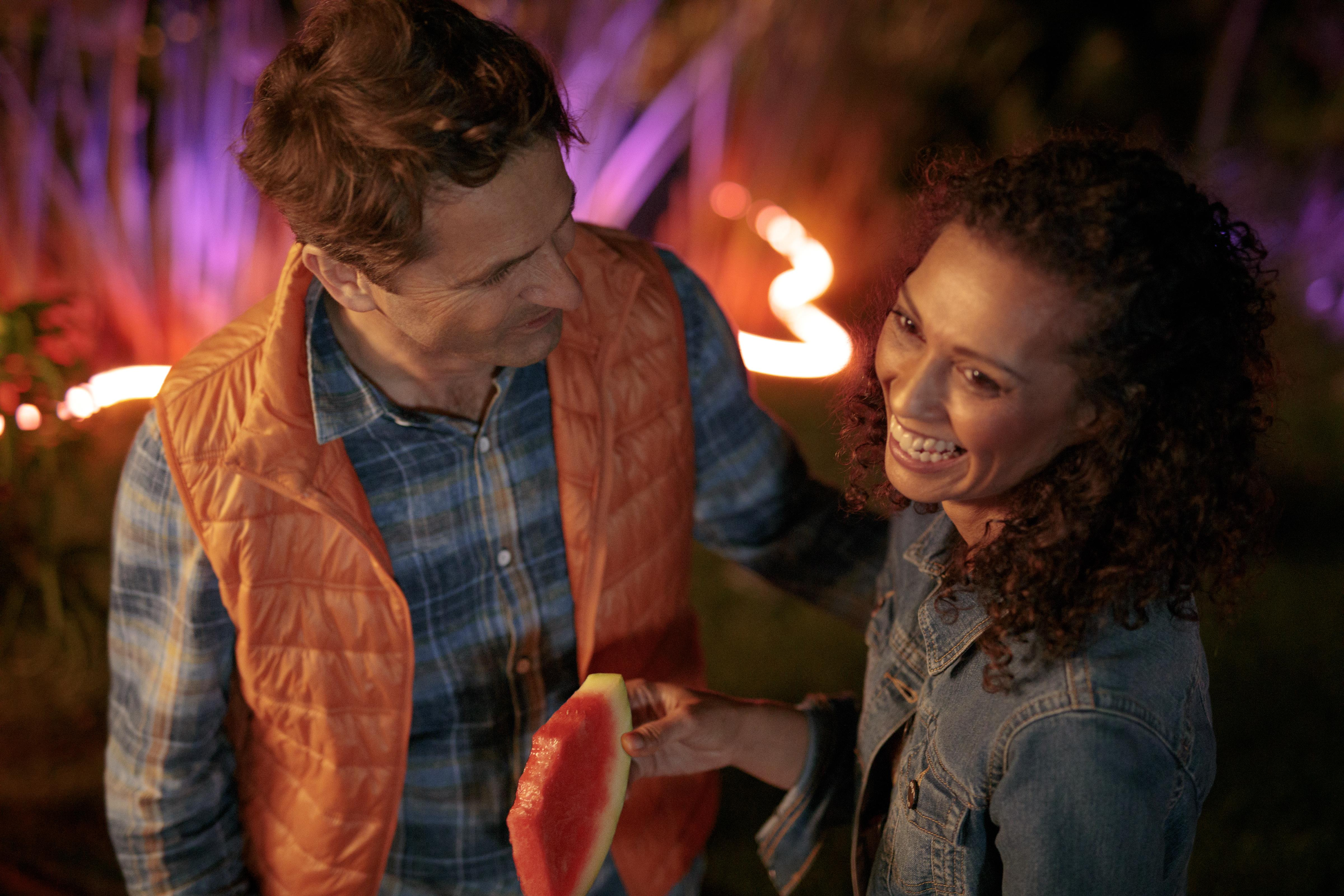 Para na zewnątrz, uśmiechnięta, trzymająca arbuza. Mężczyzna w pomarańczowej kamizelce, kobieta w kurtce dżinsowej, noc.