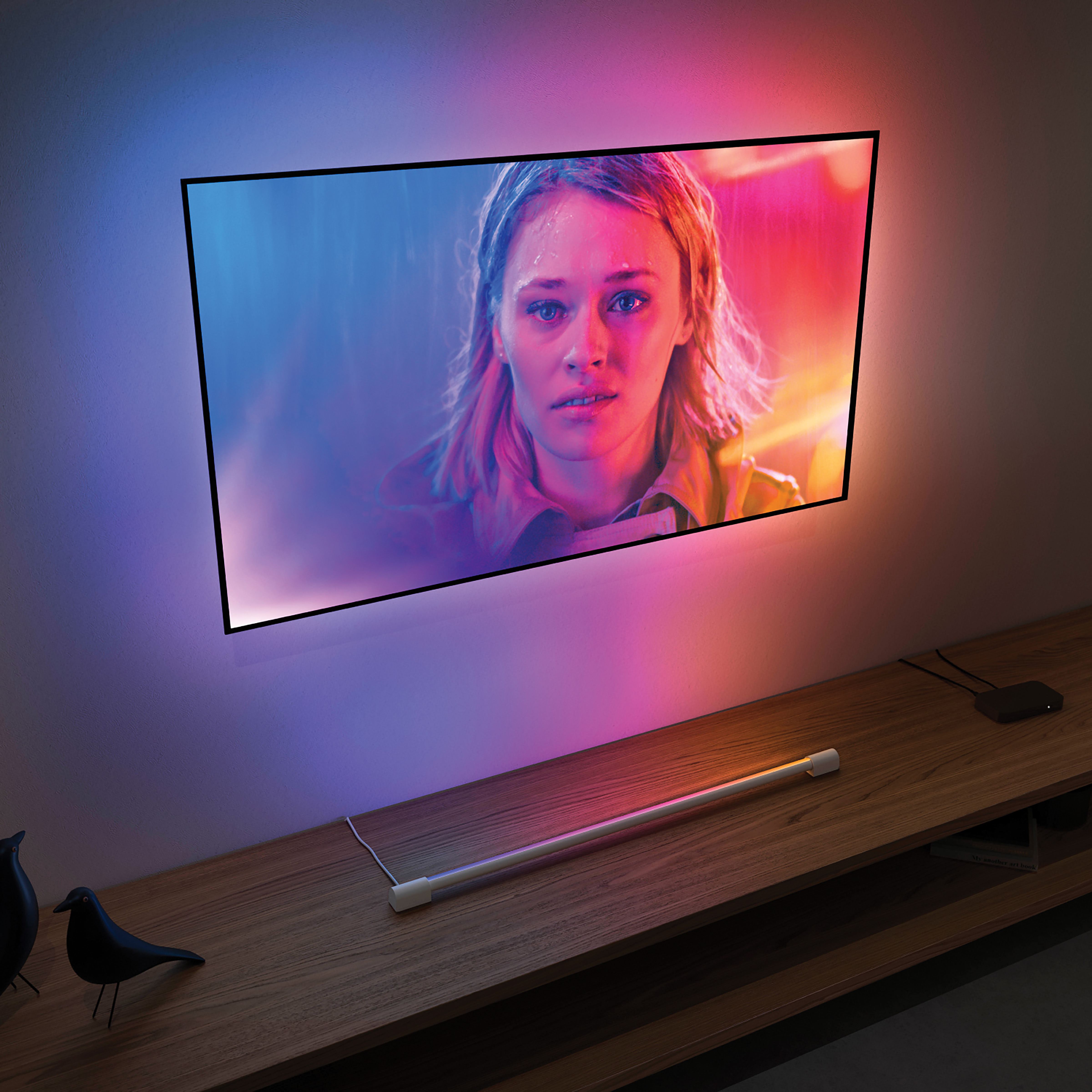 TV-Bildschirm mit dem Gesicht einer Frau, beleuchtet von bunten Lichtern an einer Wand, darunter ein Holzregal.