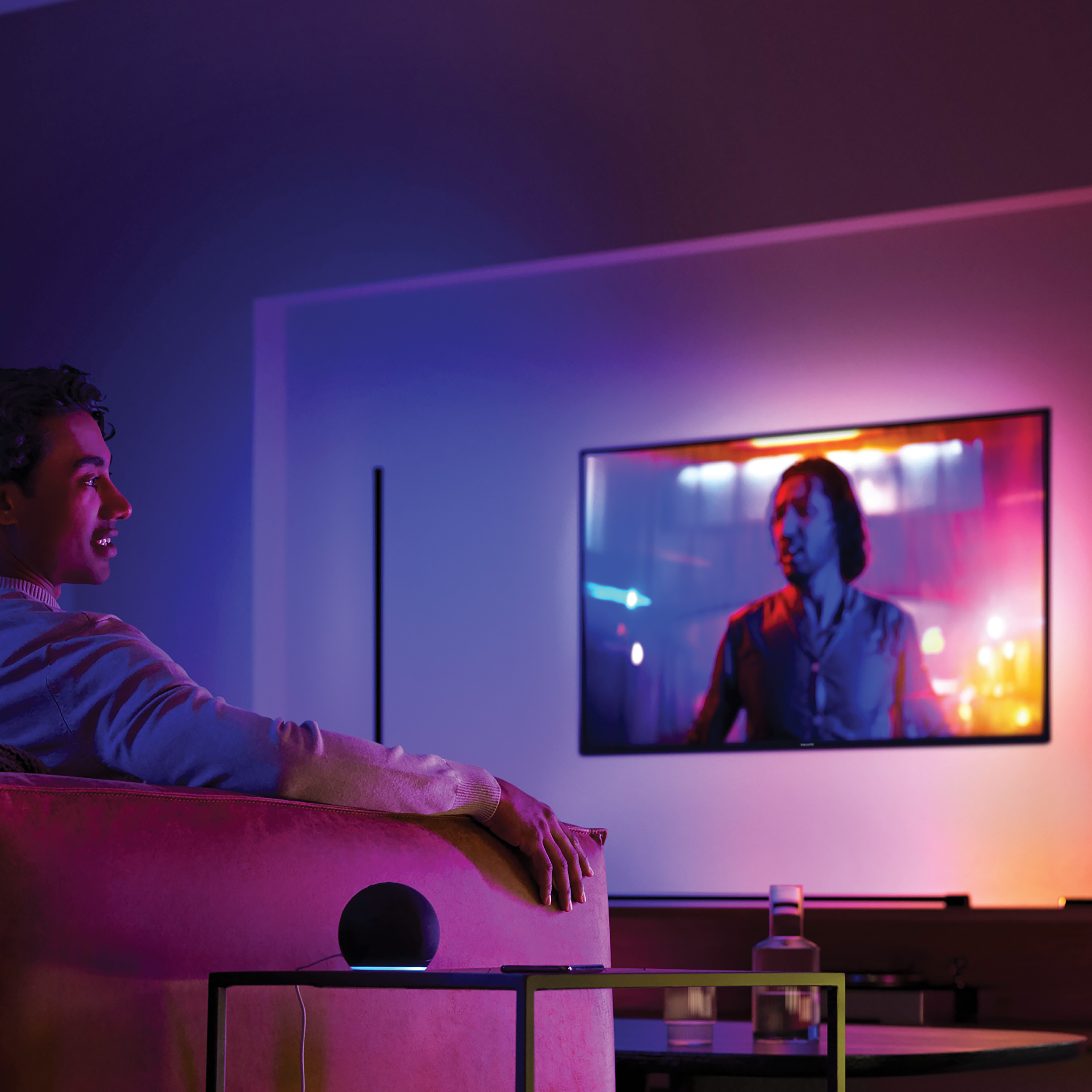 Mann auf Sofa schaut Fernsehen mit farbiger Ambientebeleuchtung und Smart Speaker.