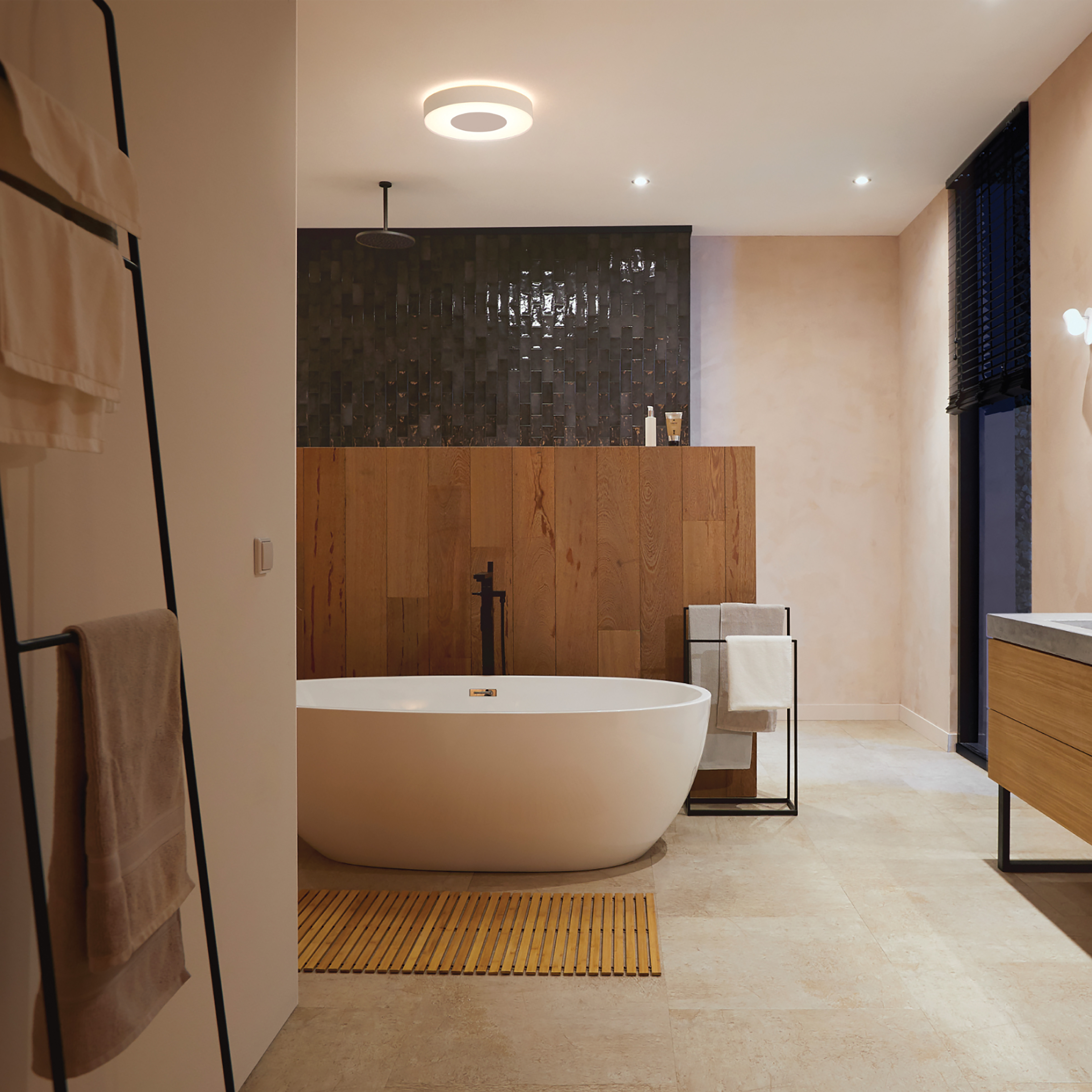 Łazienka z białą wanną, drewnianą ścianą, czarnymi kafelkami i wieszakiem na ręczniki. Beżowa podłoga i oświetlenie.