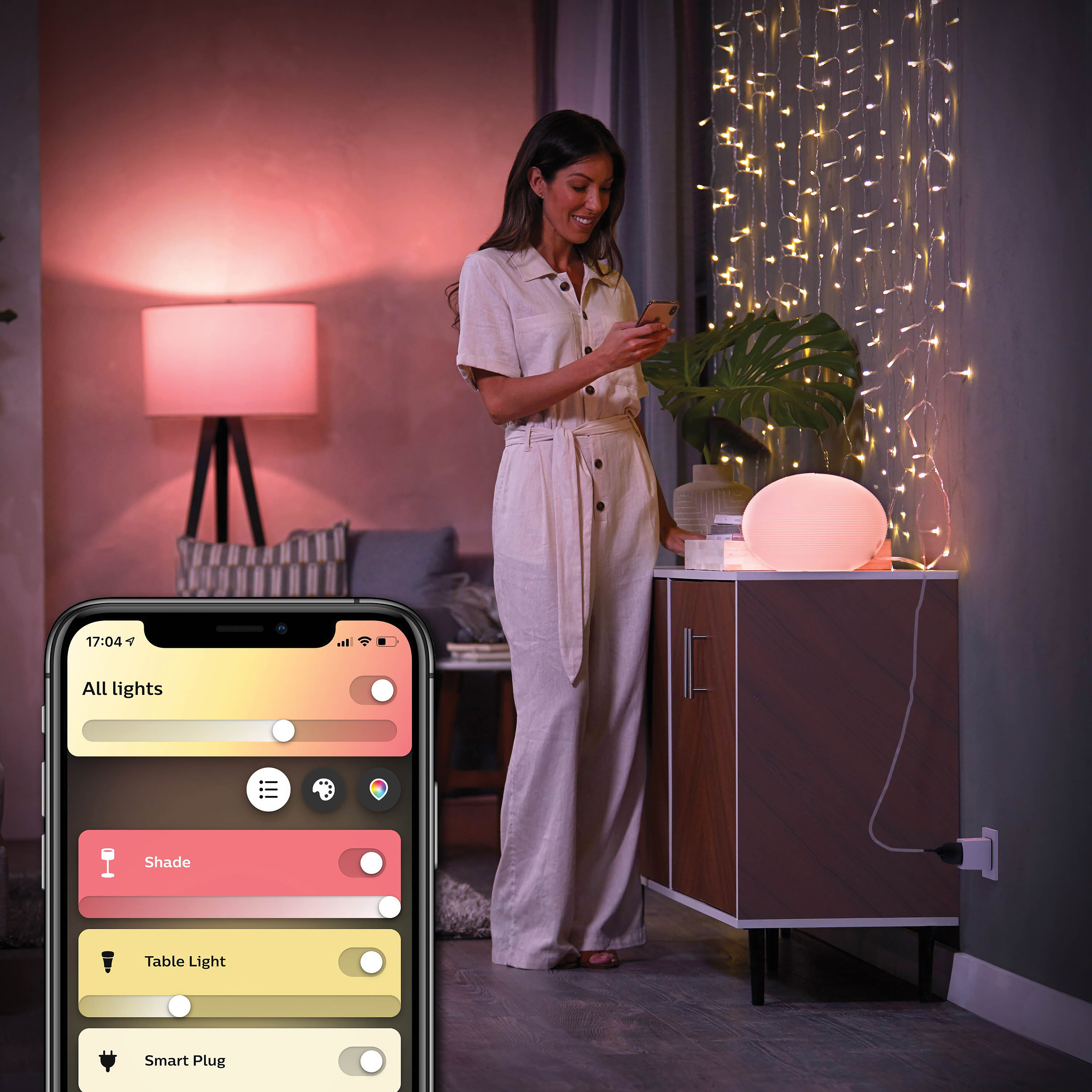 A woman is using her phone to control smart lights. A lamp and string lights are visible. The app interface is on the phone.