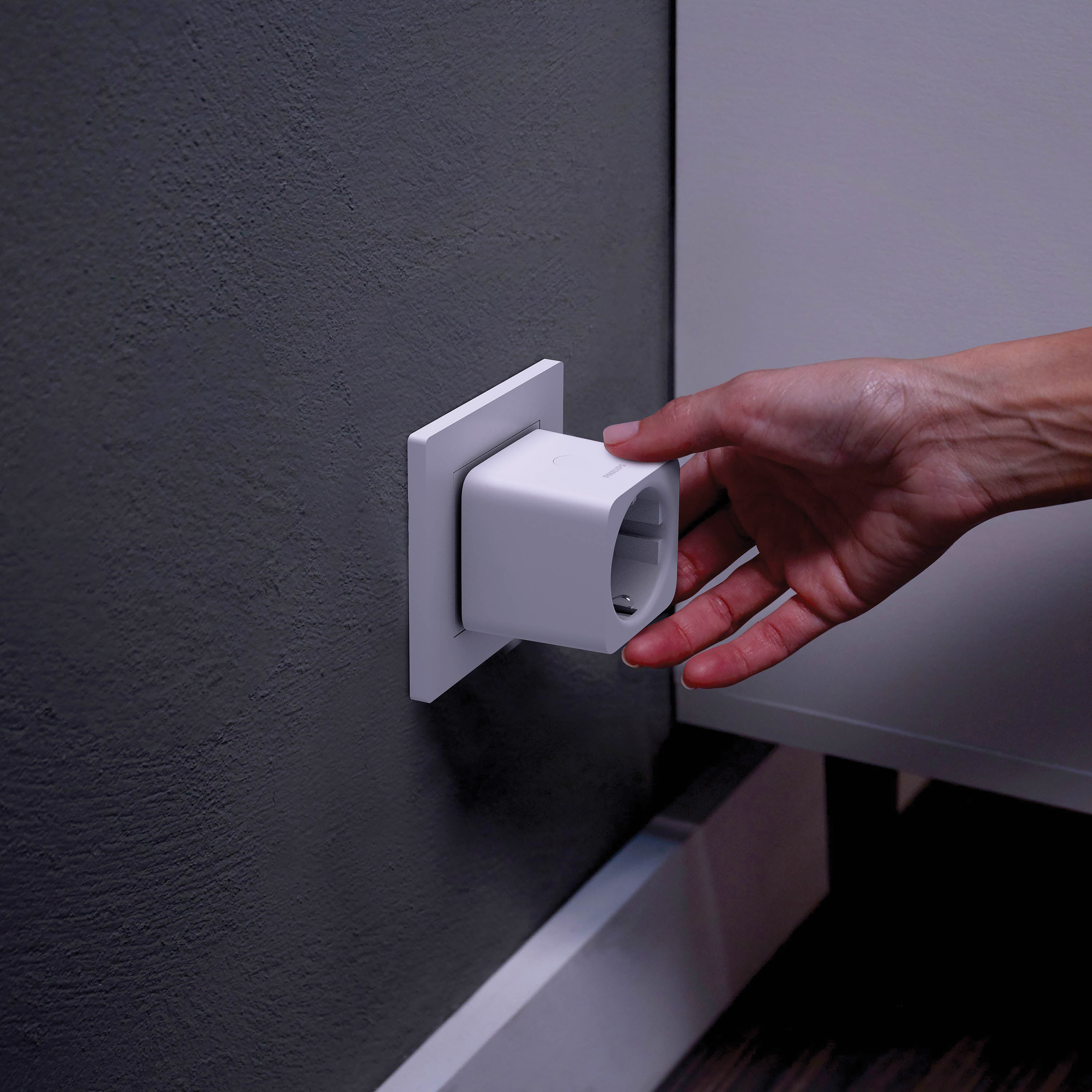 A hand plugging a white smart plug into a wall socket on a dark grey wall.