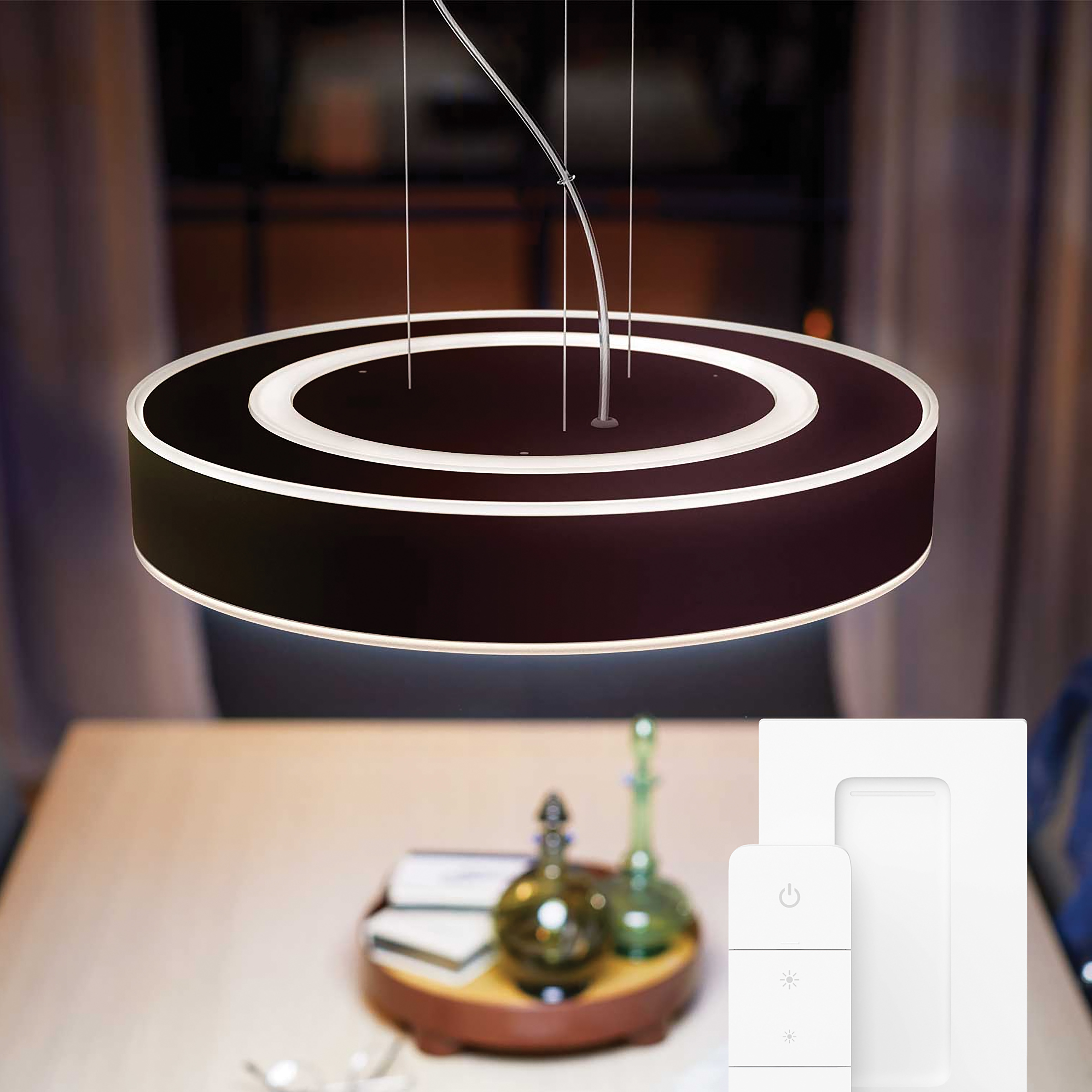 Okrągła lampa wisząca, ciemnobrązowa z białymi pierścieniami wewnętrznymi. Widoczny stół, dekoracje i ściana.