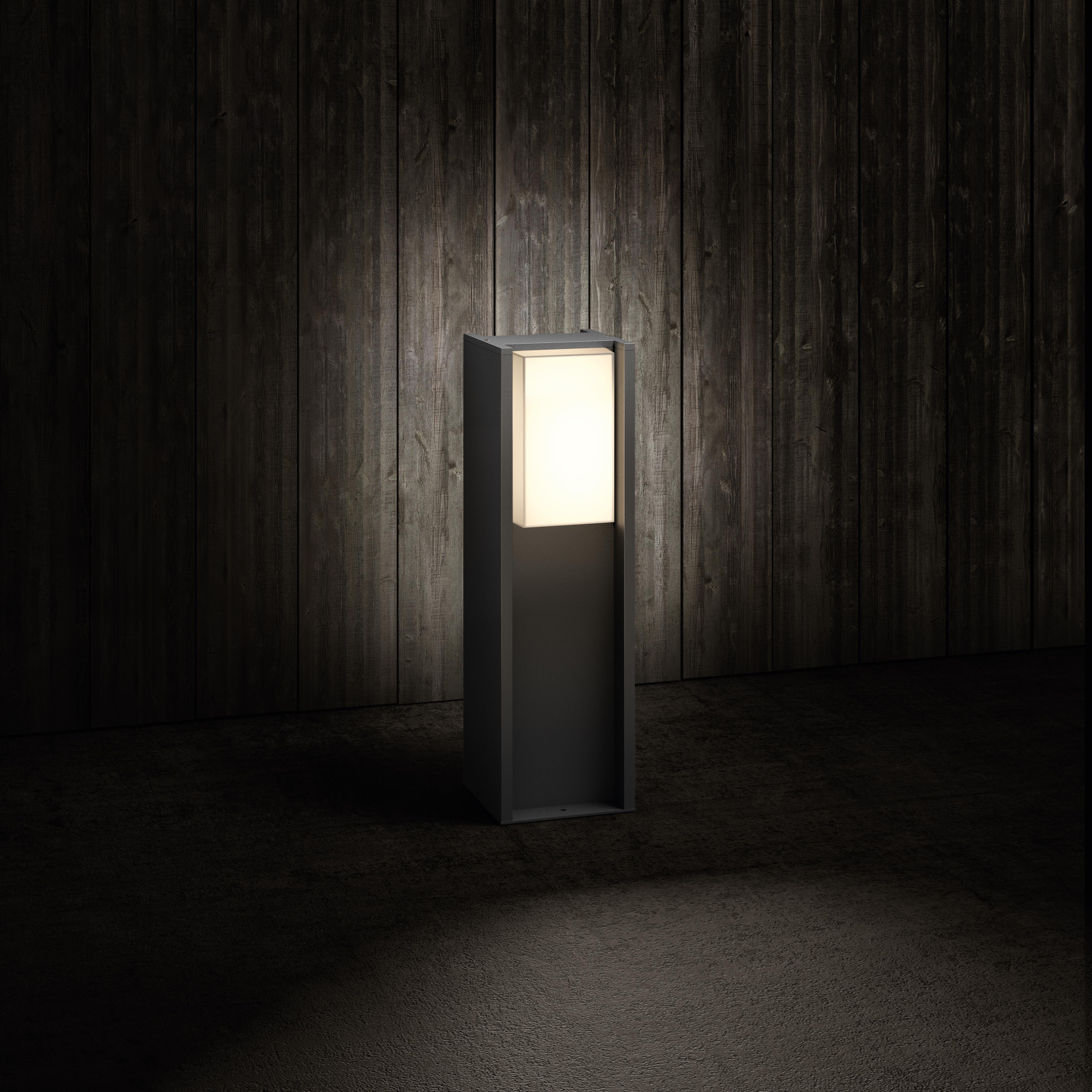 Ciemnoszara oprawa oświetleniowa zewnętrzna na tle ciemnego drewna. Światło jest prostokątne i świeci.