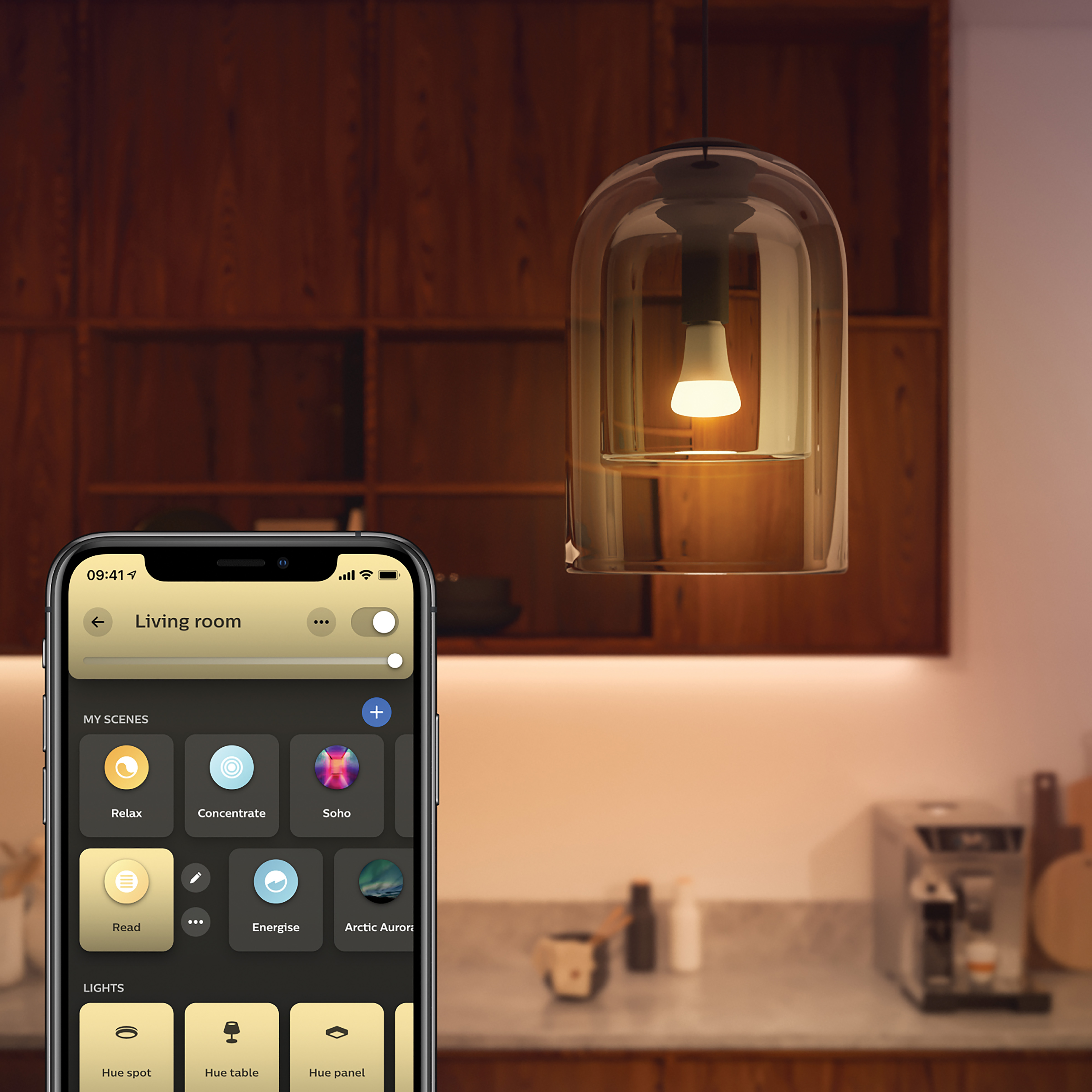 Smartfon i lampa wisząca. Widoczna drewniana szafka. Ekran telefonu pokazuje sceny.