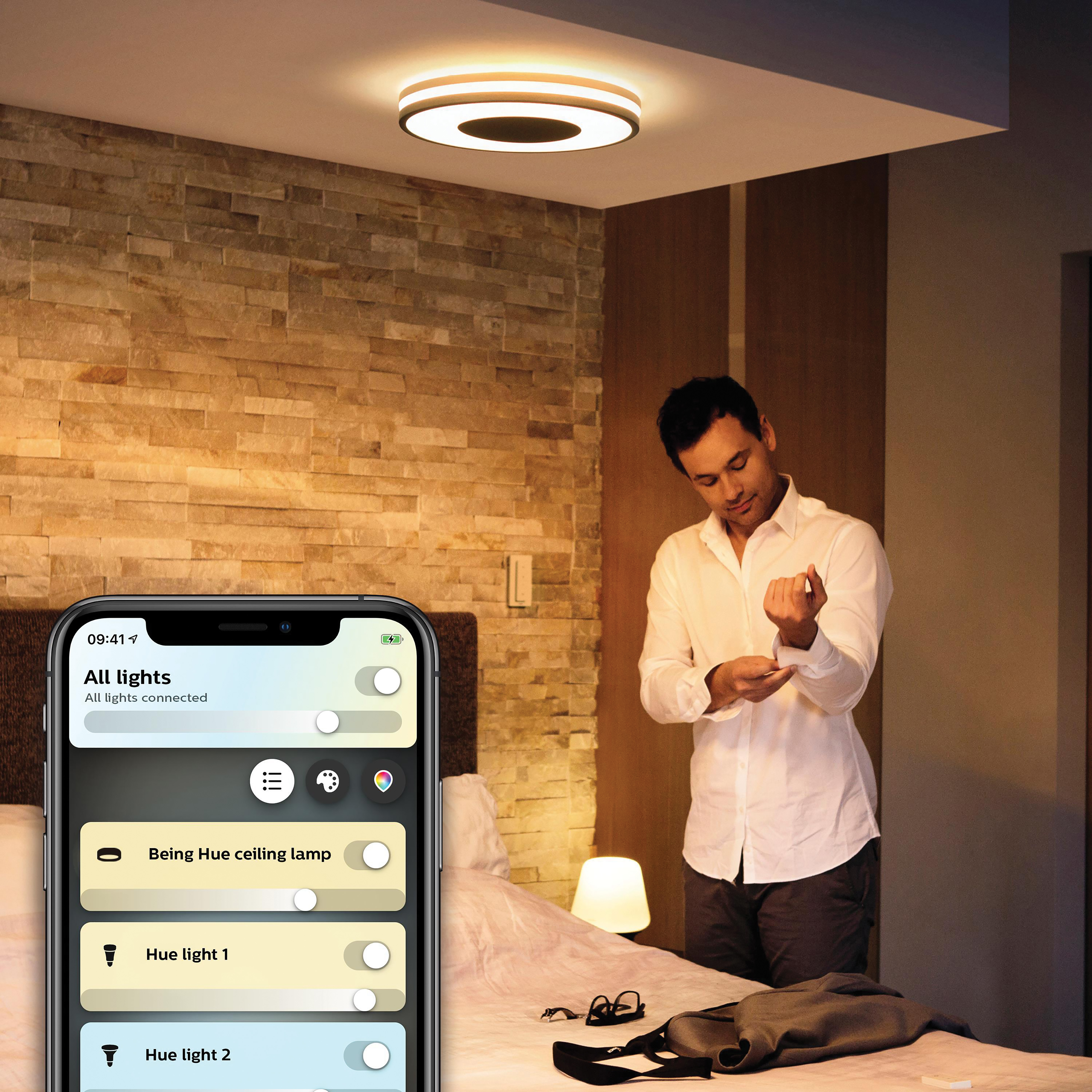 Homme ajustant chemise dans chambre. Téléphone montre appli domotique, lumière au plafond.