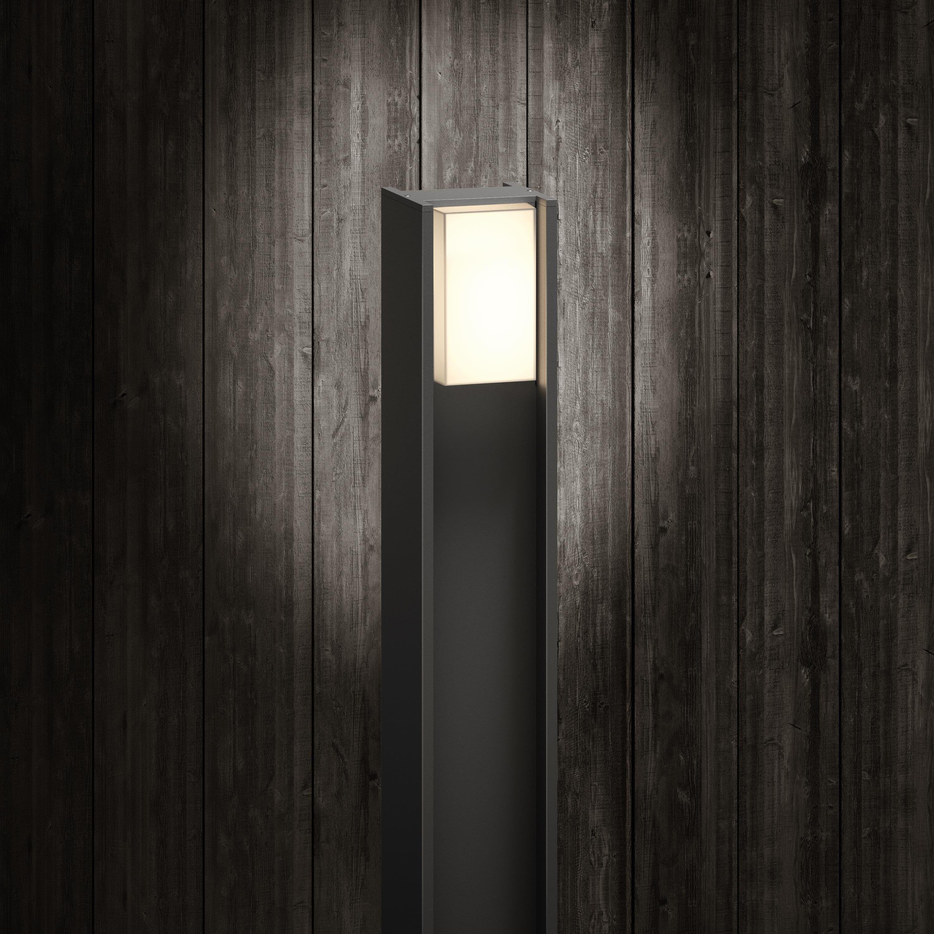 Wysoka, czarna, zewnętrzna oprawa oświetleniowa z jasnym prostokątnym światłem. Ciemne drewniane tło. Pionowa konstrukcja.
