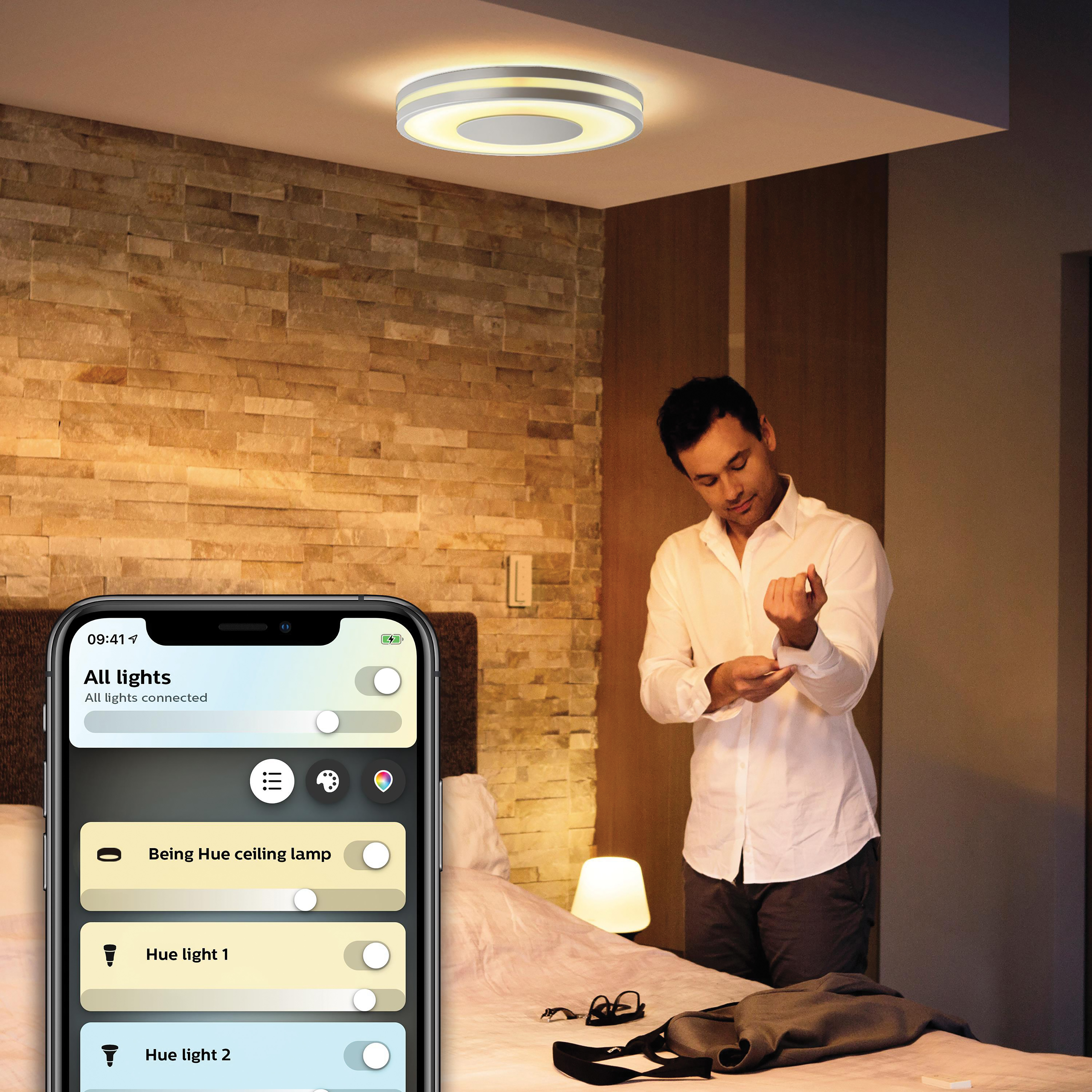 Mann in Schlafzimmer, nutzt Smart-Home-App am Telefon. Deckenleuchte sichtbar.