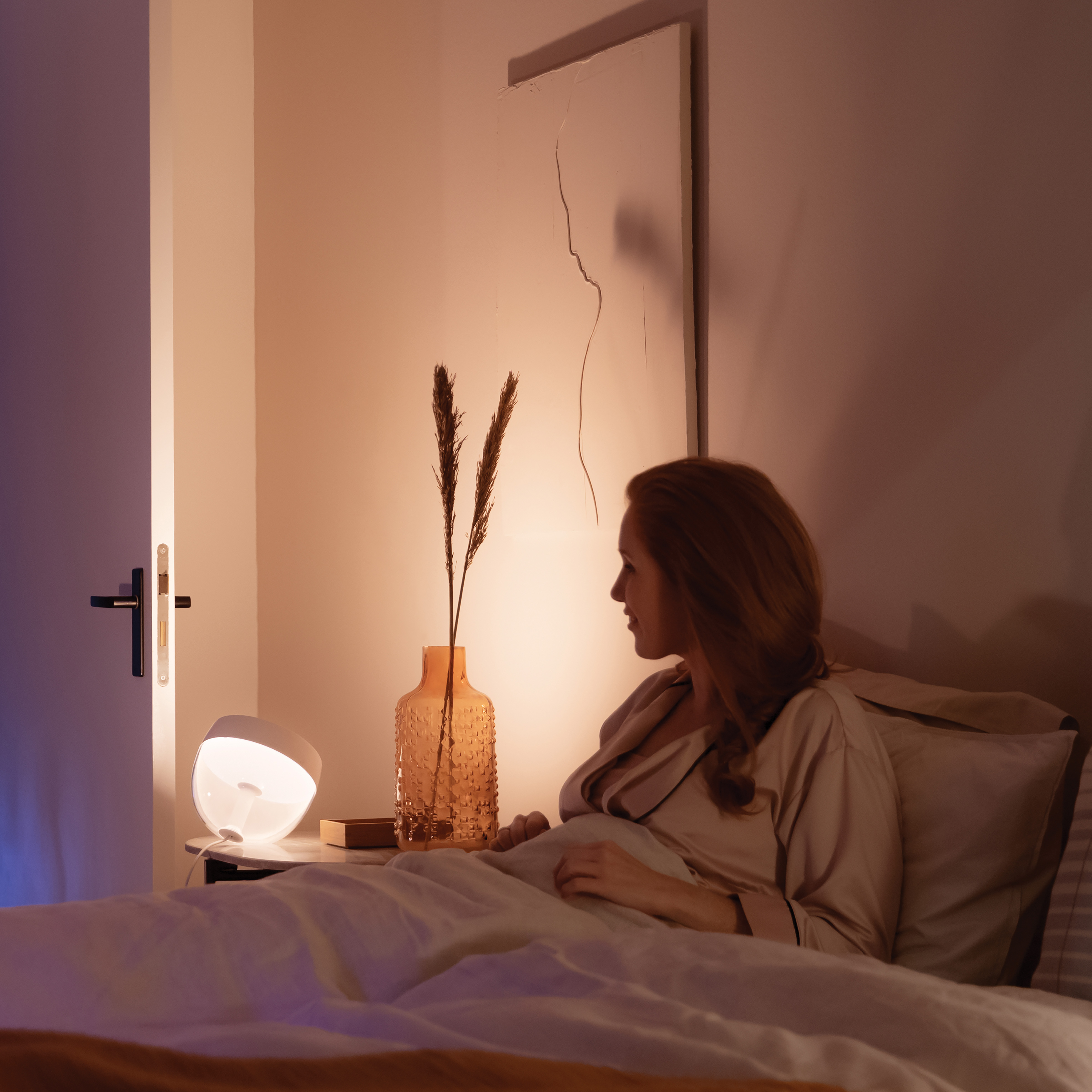 Frau im Bett, die eine Lampe auf einem Nachttisch betrachtet. Vase mit Trockenblumen daneben.