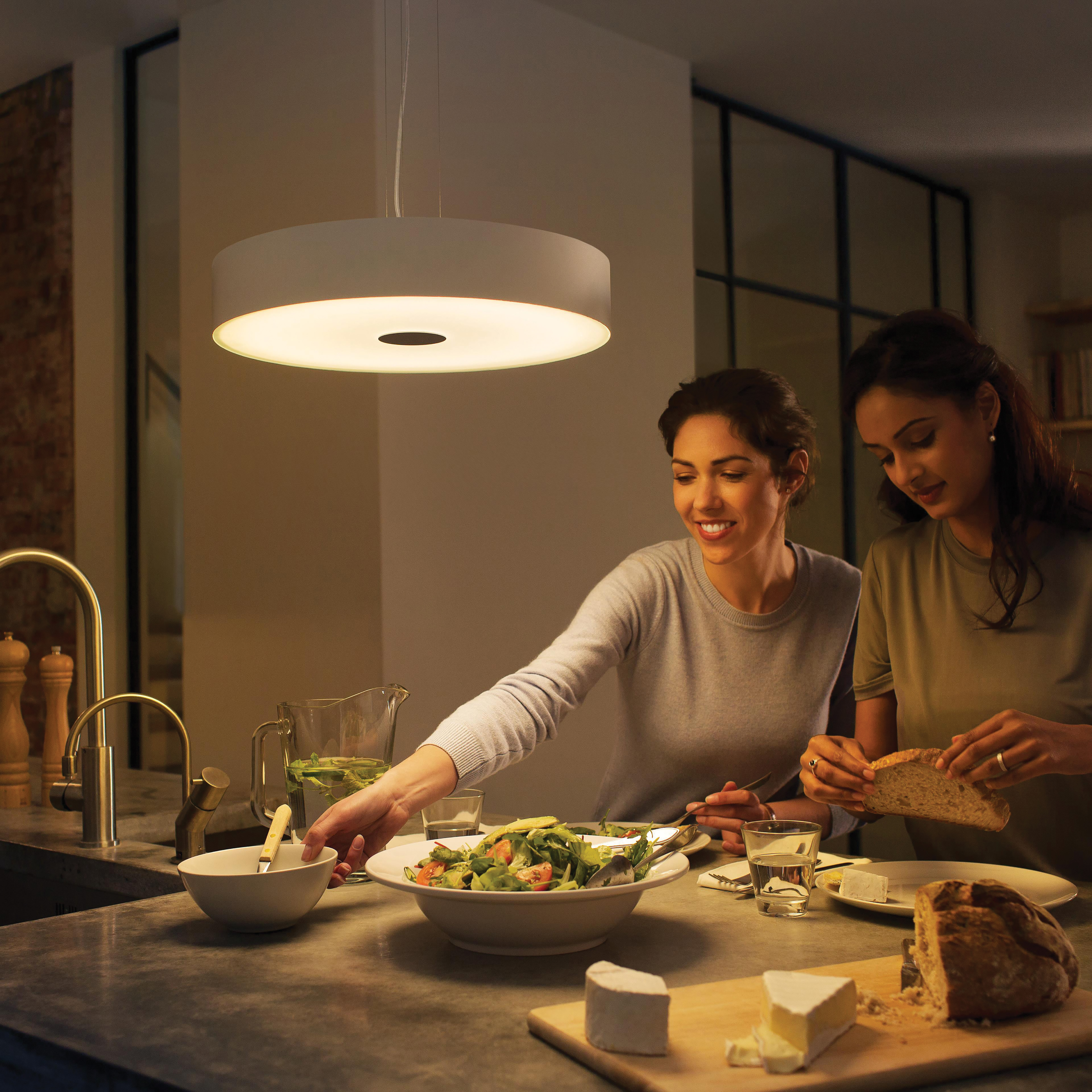 Deux femmes mangeant dans une cuisine sous une lumière circulaire. Table avec nourriture, pain, vin.